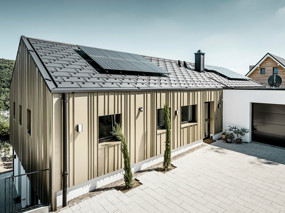 Architektonisch ansprechendes Einfamilienhaus, harmonisch in die Hügellandschaft eingebettet, mit einem hochwertigen PREFA Dachsystem in Form der Dachplatte R.16 in elegantem P.10 Anthrazit. Die Fassade zeichnet sich durch eine geschmackvolle Winkelfalzkonstruktion aus Falzonal in Lichtbronze aus, die das Gebäude mit einem warmen, metallischen Glanz umhüllt. Das Haus verfügt zudem über eine umweltfreundliche Komponente in Form von Solarmodulen, die diskret auf dem Dach integriert sind. Die PV-Anlage sowie die Gebäudehülle aus langlebigem Aluminium betonen das Engagement für nachhaltiges Bauen. Eine minimalistische Garage fügt sich nahtlos an das Hauptgebäude an, während die gepflasterte Einfahrt und sorgfältig platzierte Grünpflanzen das Bild eines modernen, ökologisch bedachten Zuhauses vervollständigen.