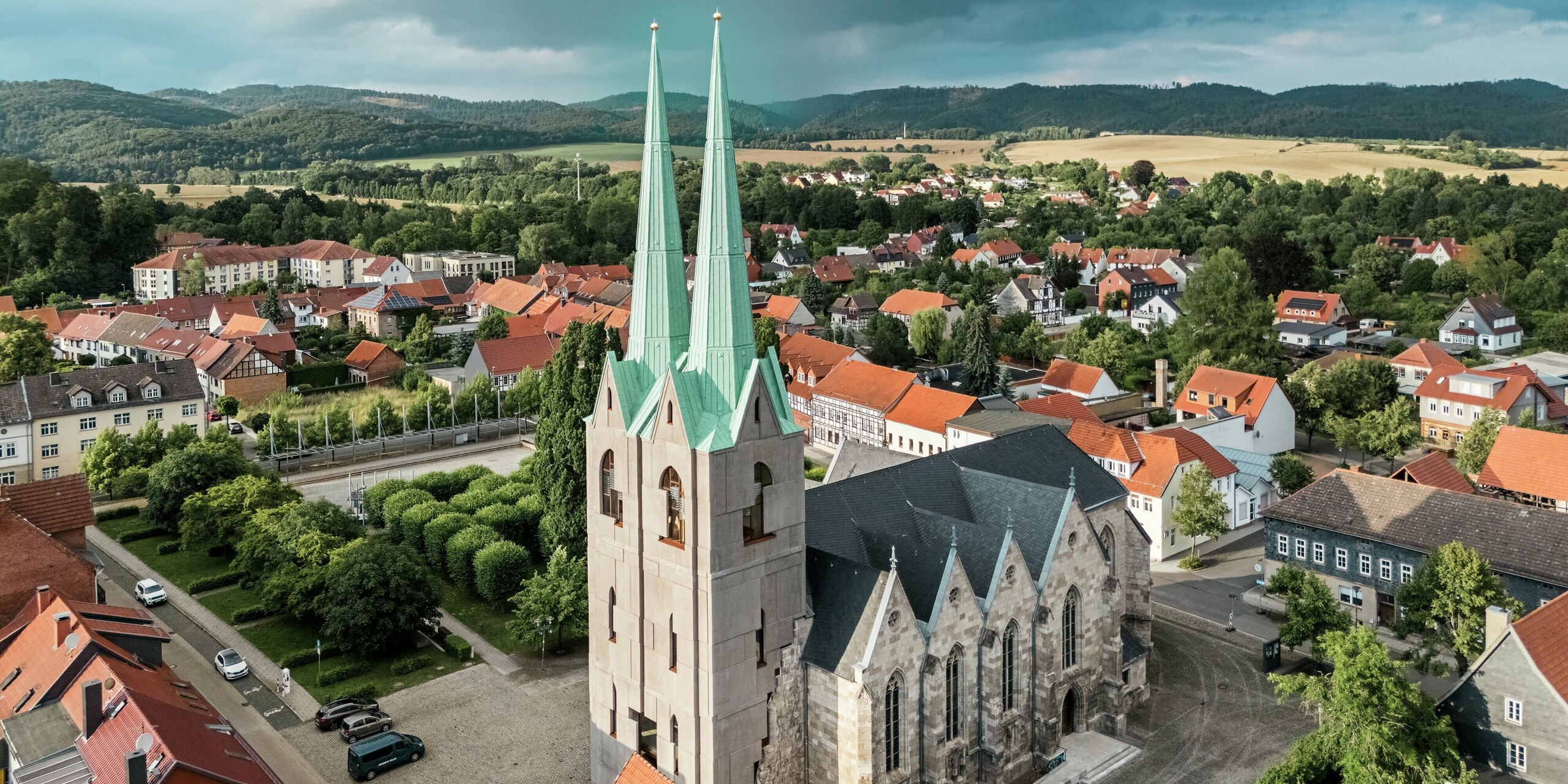 Vue aérienne de l'église Saint-Jean d'Ellrich, rénovée, avec ses deux tours jumelles impressionnantes, recouvertes de PREFALZ vert de gris P.10. L'élégante toiture en aluminium se distingue nettement des toits rouges environnants et confère à cet ensemble historique une touche de modernité. PREFALZ offre une résistance maximale aux intempéries, une grande durabilité et ne nécessite aucun entretien.