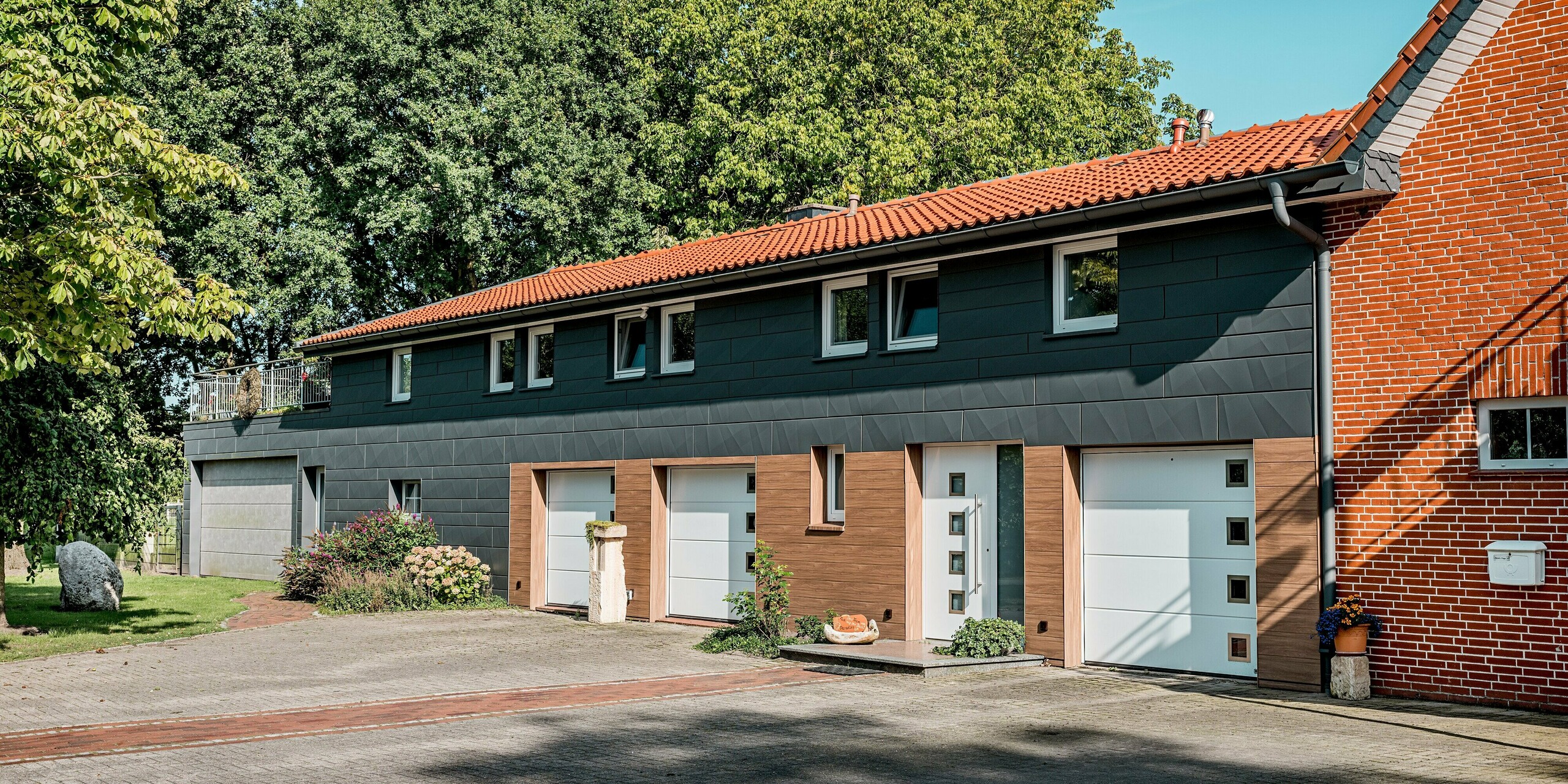 Modern saniertes Einfamilienhaus mit markanter PREFA Aluminiumfassade in horizontaler Gliederung. Eyecatcher sind die Kombination aus PREFA Siding in warmer Walnussbraun-Holzoptik im Sockel- und Eingangsbereich sowie Siding.X in P.10 Dunkelgrau im Obergeschoss. Die präzisen Kantungen der dunklen Paneele erzeugen spannende Licht- und Schattenspiele und verleihen der Fassade eine zeitgemäße Eleganz. Weiße Garagentore und klar gefasste Fenster setzen bewusste Kontraste. Die langlebigen, witterungsbeständigen und pflegeleichten PREFA Aluminiumpaneele eignen sich ideal für nachhaltige Sanierungen und verbinden modernes Design mit hoher Funktionalität.
