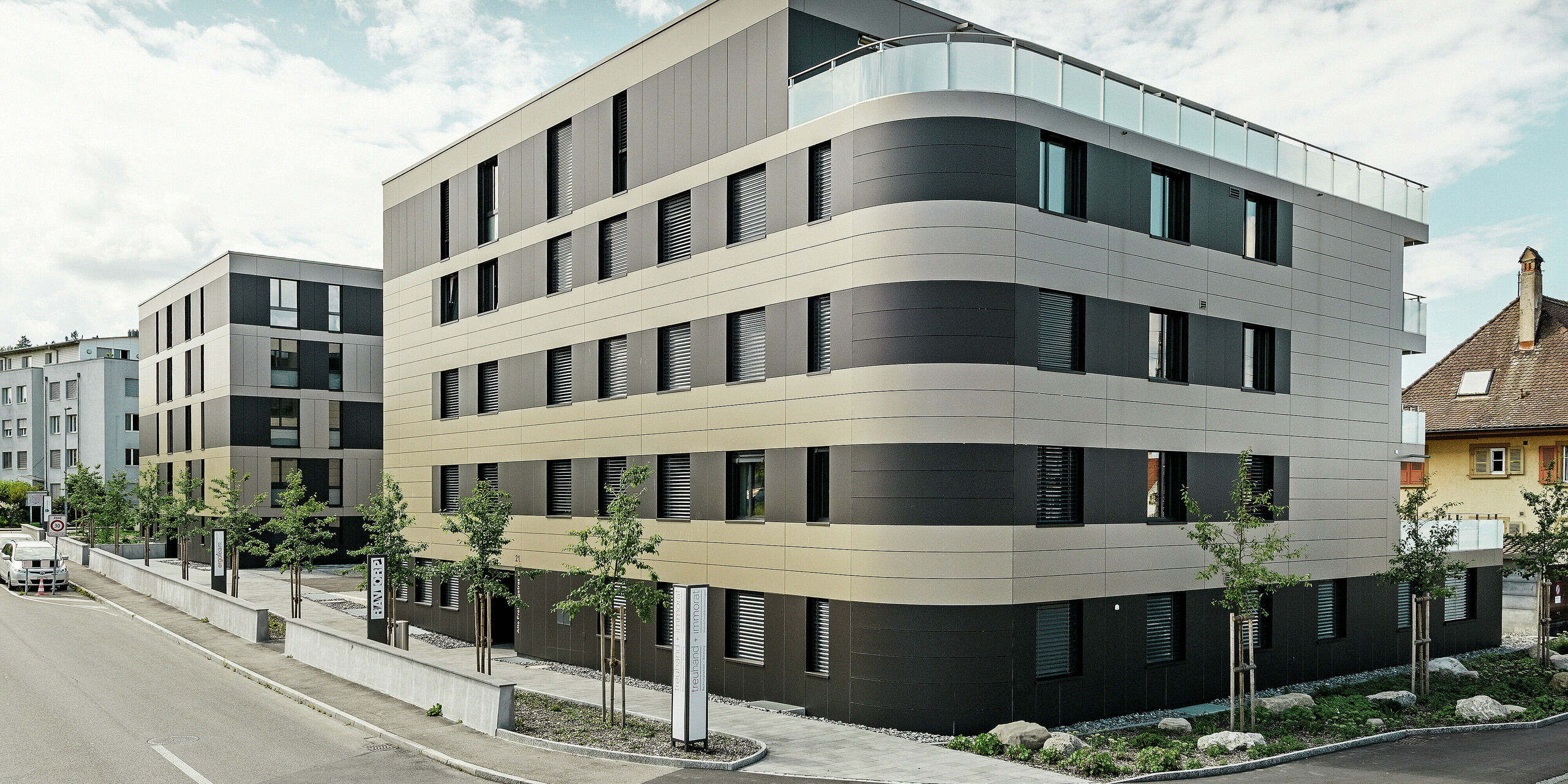 In der kubischen Wohnanlage von Murten (CH) sorgen die PREFA Aluminium Verbundplatten in Bronze und Schwarzgrau für eine optische Aufwertung und gleichzeitig für eine robuste und langlebige Fassade. Die Kombination dieser Farbtöne bietet ästhetische Akzente und schützt das Gebäude langfristig vor Wettereinflüssen. An der Gebäudeecke zur Straße hin ist eine Rundung zu erkennen, was das Rollbiegen der PREFABOND Aluminium Verbundplatten demonstrieren soll.