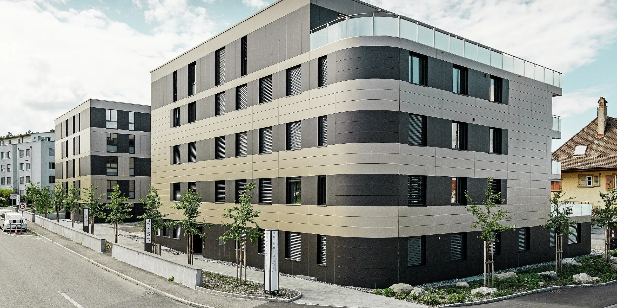 In der kubischen Wohnanlage von Murten (CH) sorgen die PREFA Aluminium Verbundplatten in Bronze und Schwarzgrau für eine optische Aufwertung und gleichzeitig für eine robuste und langlebige Fassade. Die Kombination dieser Farbtöne bietet ästhetische Akzente und schützt das Gebäude langfristig vor Wettereinflüssen. An der Gebäudeecke zur Straße hin ist eine Rundung zu erkennen, was das Rollbiegen der PREFABOND Aluminium Verbundplatten demonstrieren soll.