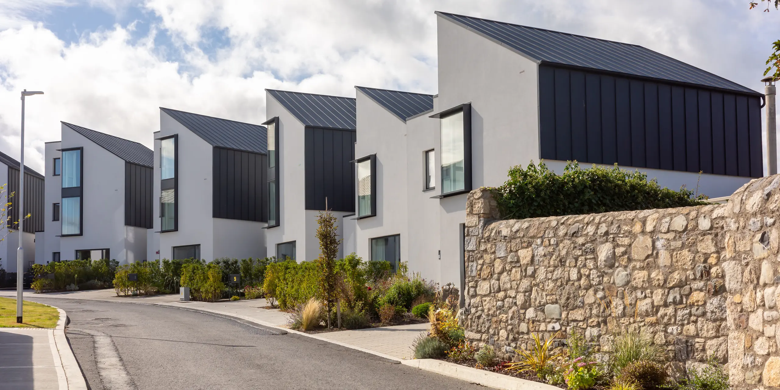 Le complexe résidentiel Watson Place se distingue par ses toitures inclinées aux formes originales et son bardage continu PREFALZ couleur P.10 anthracite. Les surfaces en aluminium foncé contrastent fortement avec les façades claires et les murs en pierre naturelle. L'aluminium PREFA, d'une grande durabilité, assure une protection efficace contre les intempéries et confère à cette architecture moderne une allure épurée et haut de gamme.