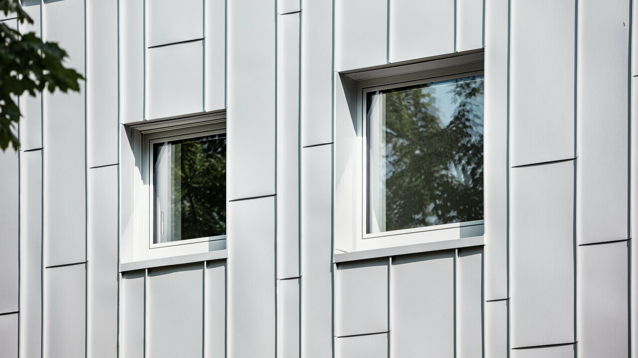 Gros plan sur la façade d'un bâtiment moderne avec des panneaux verticaux en aluminium prépliés argentés. Deux fenêtres rectangulaires aux cadres sombres reflètent le vert des arbres et rompent le motif structuré de la façade.