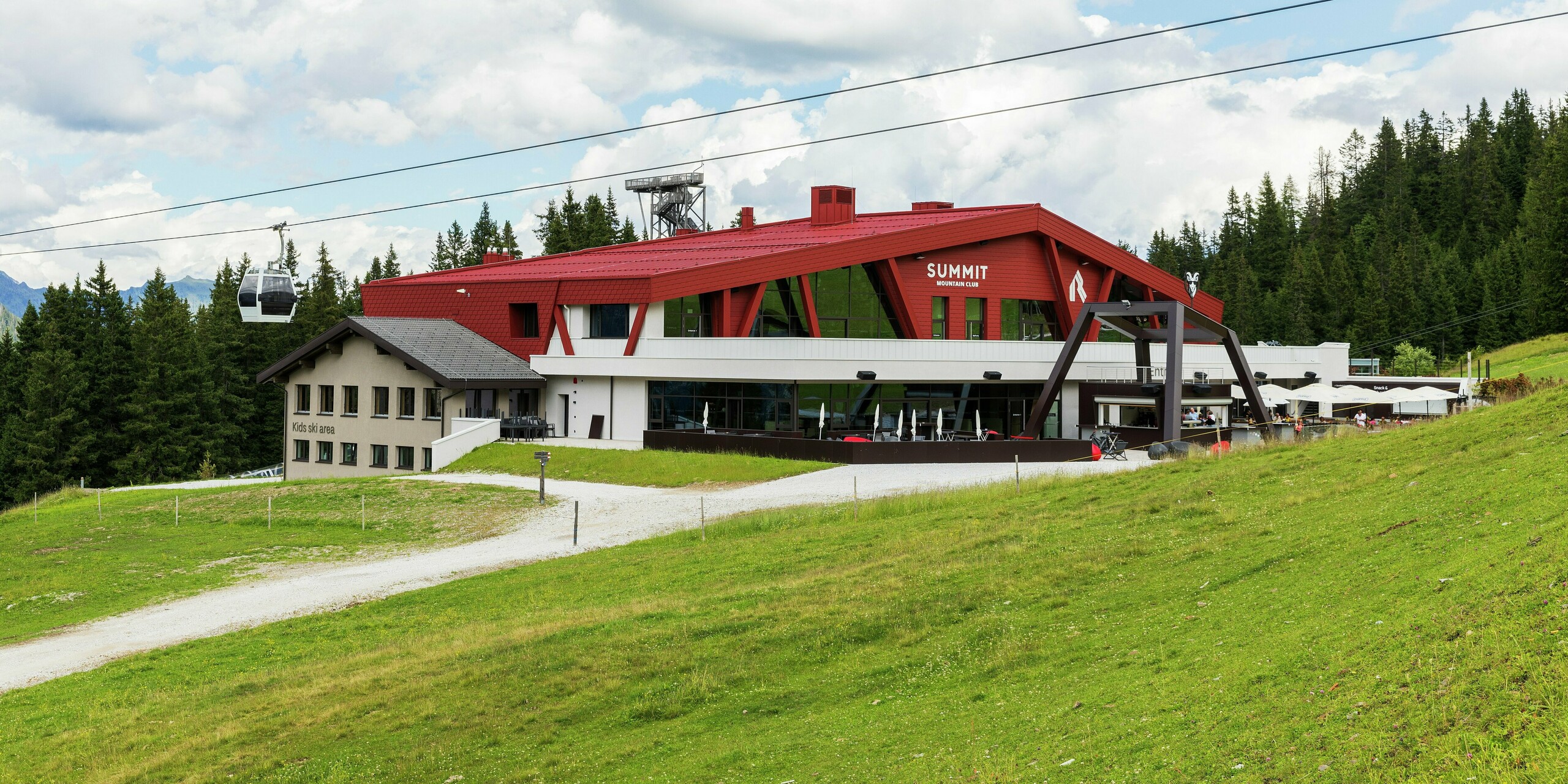 Le Summit Mountain Club de Ratschings se distingue par son toit remarquable et sa façade couleur P.10 rouge oxyde. Son architecture moderne s'intègre harmonieusement au paysage alpin. La structure, audacieuse et dotée de larges baies vitrées, attire immédiatement le regard. La route d'accès est visible au premier plan. Revêtu de bardeaux PREFA et de panneaux à joints debout PREFALZ, le bâtiment offre des solutions à la fois esthétiques et fonctionnelles, parfaitement adaptées aux rigueurs du climat montagnard.