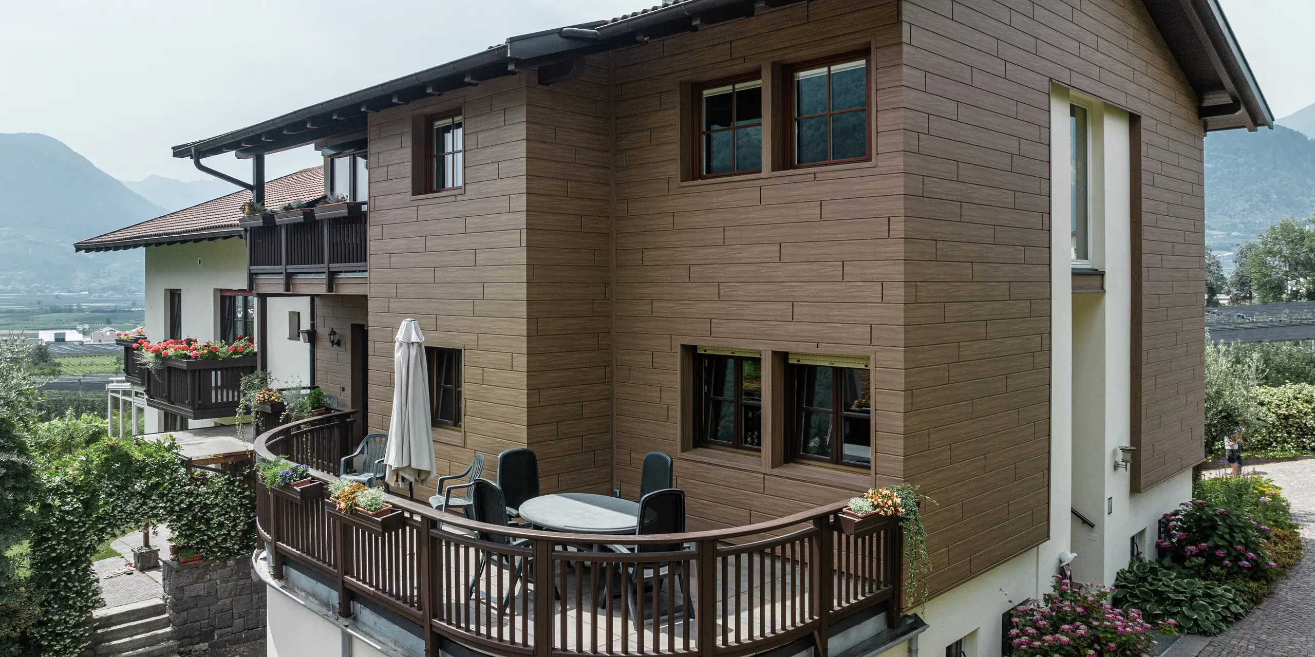 Sanierte Ferienwohnungen Prechtlhof in Meran, Südtirol, mit PREFA Sidings in Walnussbraun. Die wartungsfreie Aluminiumfassade in Holzoptik verbindet moderne Ästhetik mit traditionellem Charme und fügt sich harmonisch in die alpine Umgebung ein. Der Rundbalkon im Vordergrund lädt zum Verweilen in der traumhaften Südtiroler Landschaft ein. 