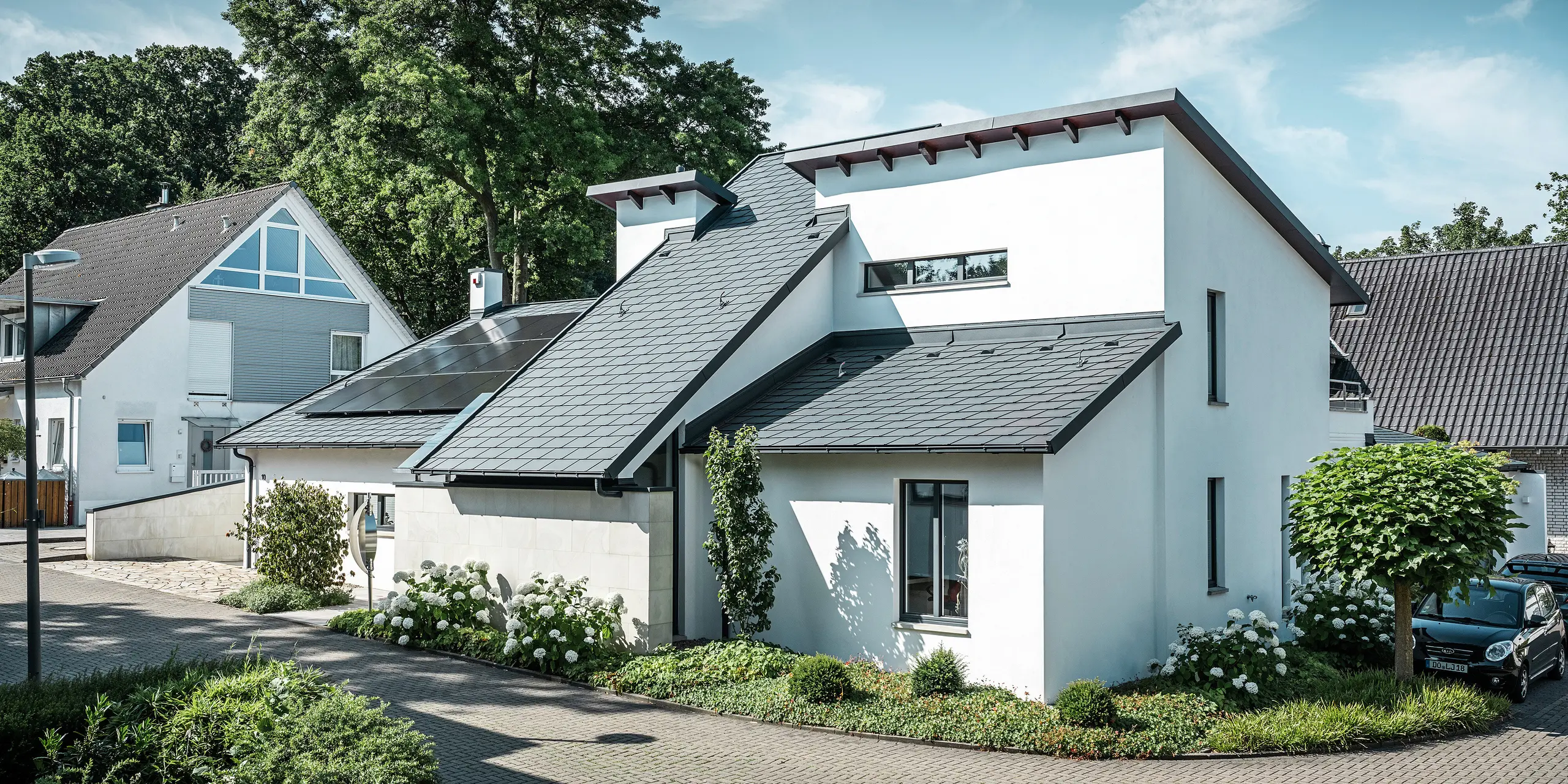 Moderna casa unifamiliare con facciata intonacata di bianco e tetto spiovente PREFA in alluminio scuro. Sul tetto sono visibili moduli fotovoltaici integrati e un sistema di smaltimento acque piovane in alluminio color antracite. L'edificio si trova in un curato complesso residenziale con giardini antistanti, vialetto pavimentato e case unifamiliari circostanti.