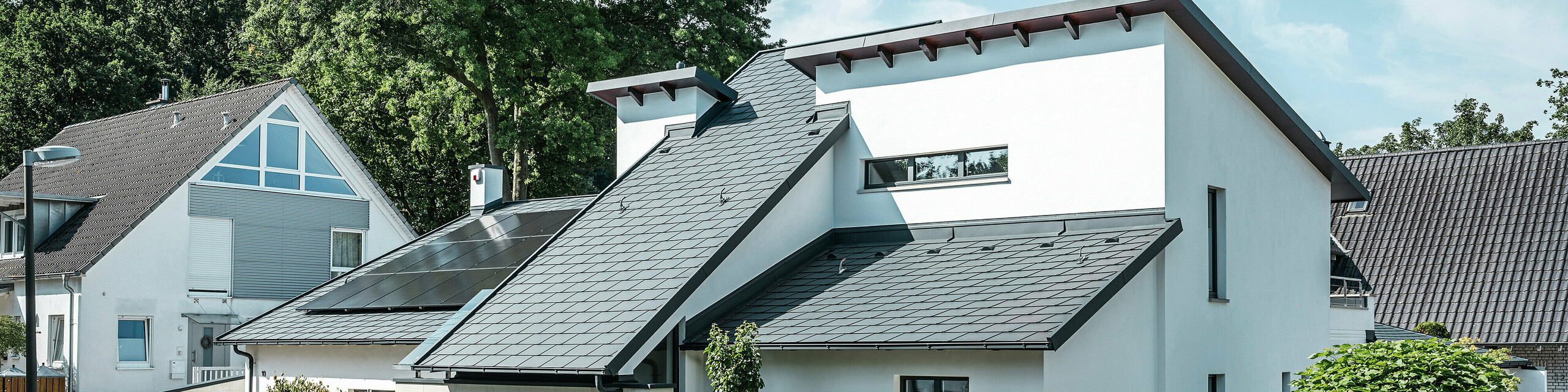 Modernes Einfamilienhaus mit weißer Putzfassade und dunklem PREFA Schrägdach aus Aluminium. Auf dem Dach sind integrierte Photovoltaikmodule und eine Aluminium-Dachentwässerung in Anthrazit zu sehen. Das Gebäude liegt in einer gepflegten Wohnsiedlung mit bepflanzten Vorgärten, gepflasterter Zufahrt und umliegenden Einfamilienhäusern.
