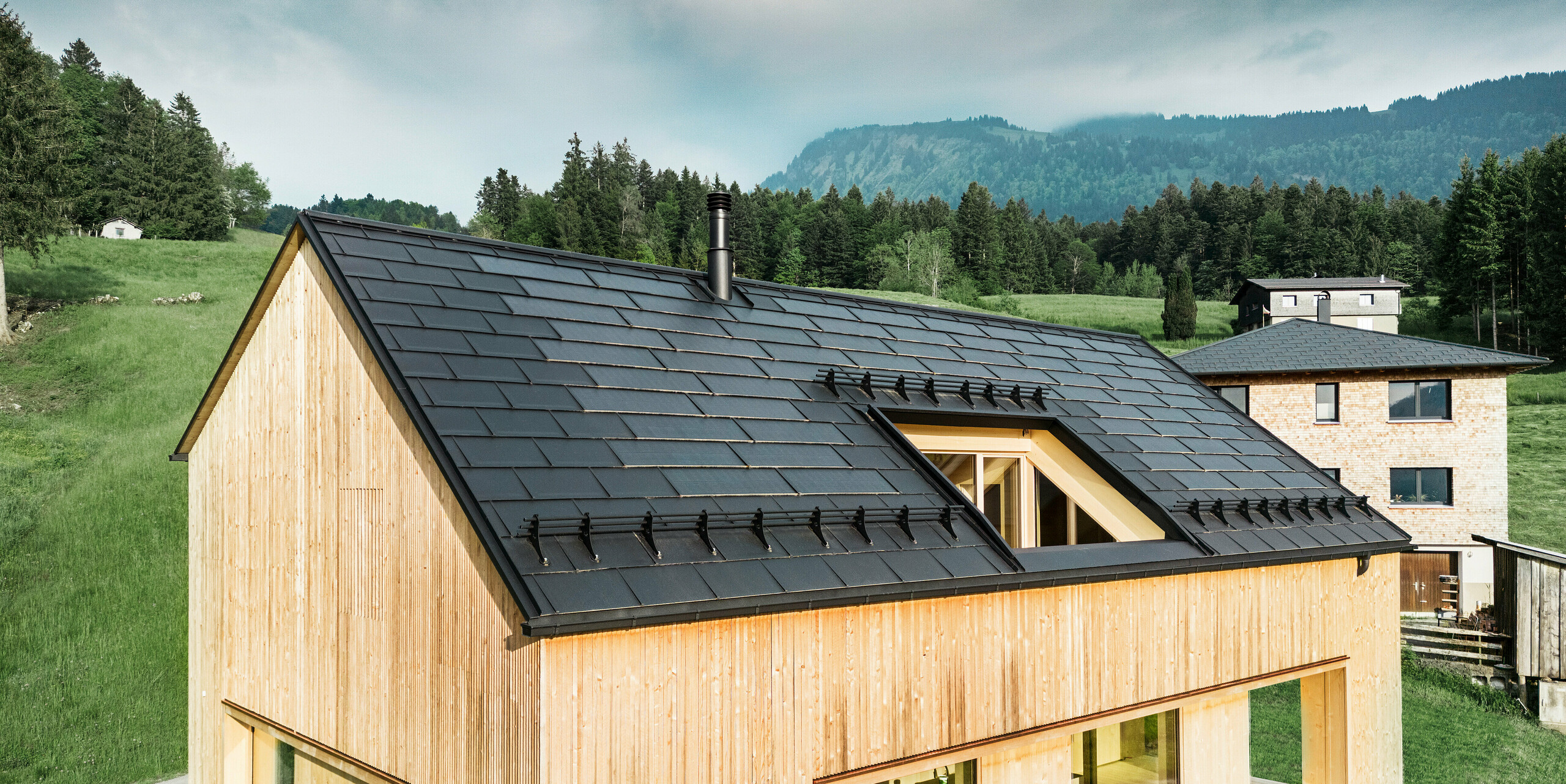 Vue de face d'une maison moderne en bois à Egg, qui se détache sur le paysage verdoyant et luxuriant avec son toit solaire PREFA sombre. Les tuiles solaires noires PREFA posés avec précision offrent non seulement une protection, mais aussi une efficacité énergétique grâce à la technologie solaire intégrée. Des R.16 PREFA traditionnels noirs ont été posés le long des bords du toit jusqu'à l'avant-toit. La cheminée classique et la grande lucarne complètent le concept durable du bâtiment, qui équilibre l'architecture moderne et les pratiques de construction respectueuses de l'environnement.
