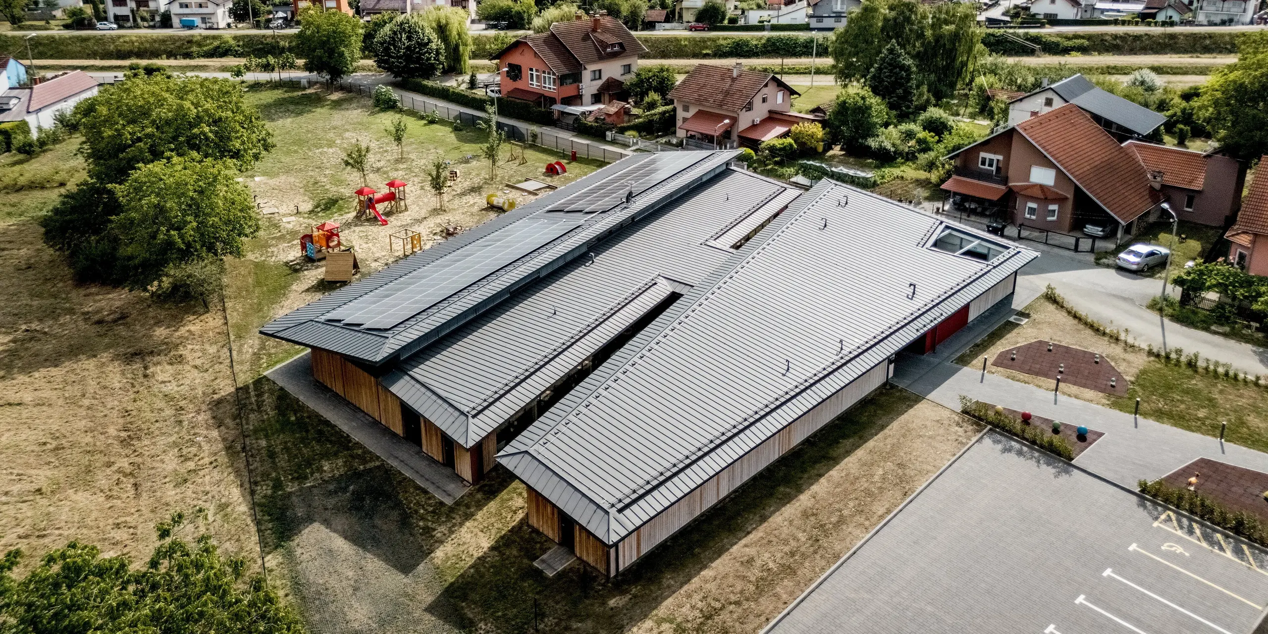 Luftaufnahme des Kindergartens Svarozic in Kroatien mit großzügiger Dachlandschaft aus PREFALZ Aluminium in P.10 Anthrazit. Das moderne Gebäude mit Holzfassade, Photovoltaikanlage und weitläufigem Spielbereich fügt sich harmonisch in die Umgebung ein. Die PREFA Dachlösung überzeugt durch langlebige Qualität, geringes Gewicht und zuverlässigen Schutz vor Witterungseinflüssen.