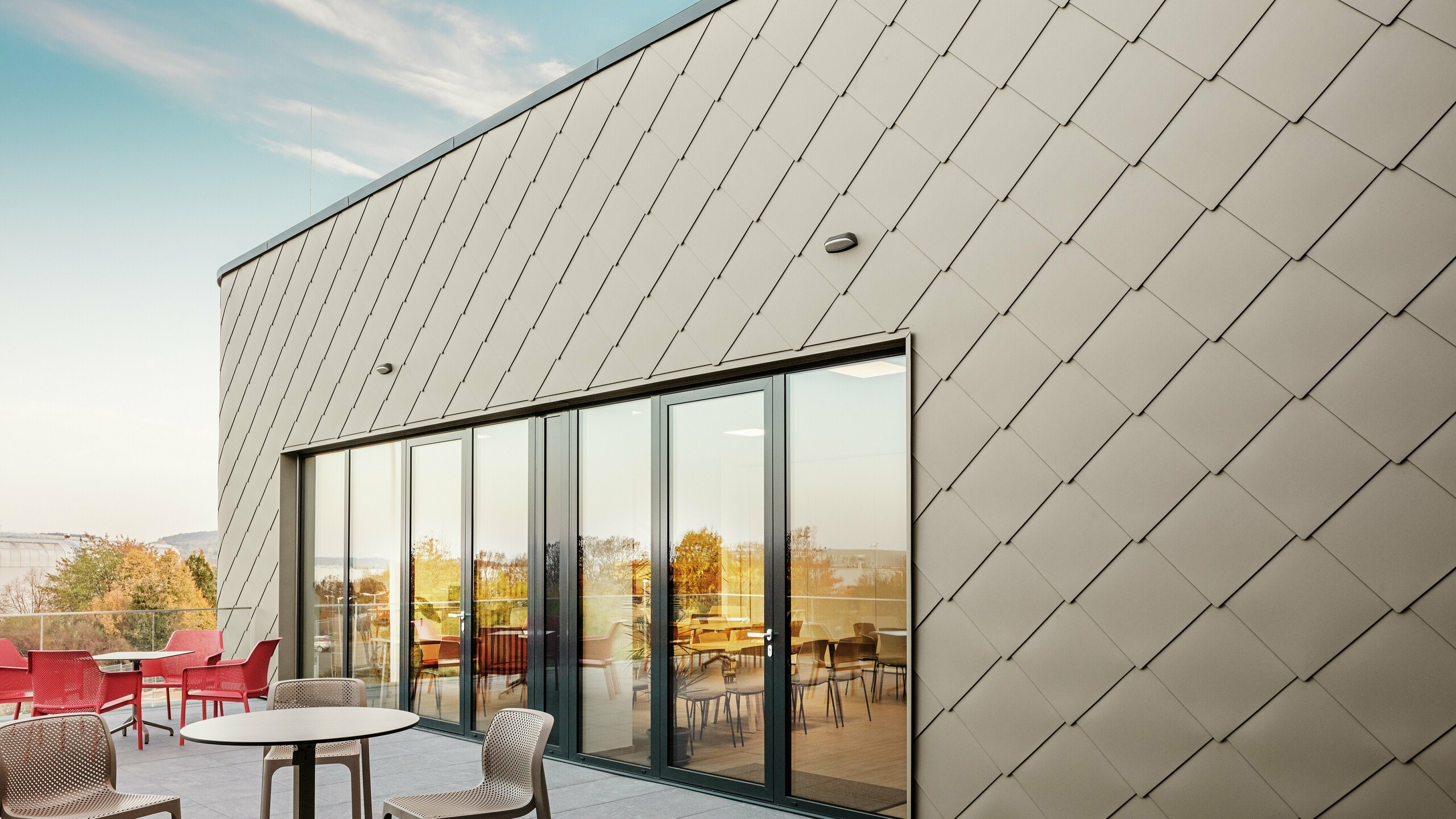 La façade du nouveau site PREFA à Törökbálint impressionne par les élégants losanges de façade PREFA 44 × 44 en bronze. La façade en aluminium posée avec précision crée une apparence moderne et dynamique tout en offrant une protection maximale contre les éléments. La généreuse façade vitrée relie les espaces intérieurs et extérieurs et crée une atmosphère lumineuse et accueillante sur la terrasse spacieuse.