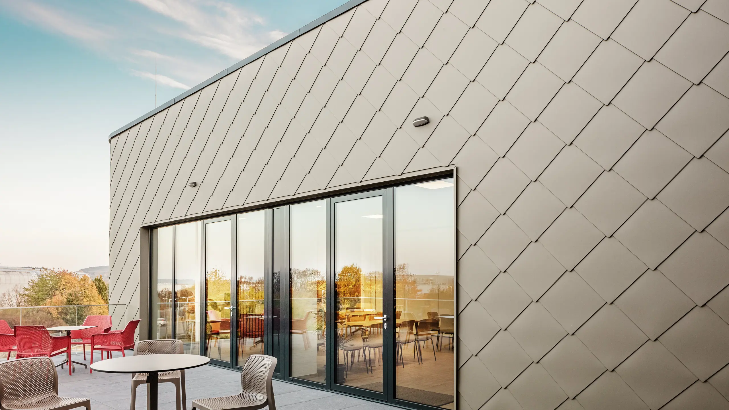La façade du nouveau site PREFA à Törökbálint impressionne par les élégants losanges de façade PREFA 44 × 44 en P.10 bronze prefa. La façade en aluminium posée avec précision crée une apparence moderne et dynamique tout en offrant une protection maximale contre les éléments. La généreuse façade vitrée relie les espaces intérieurs et extérieurs et crée une atmosphère lumineuse et accueillante sur la terrasse spacieuse.