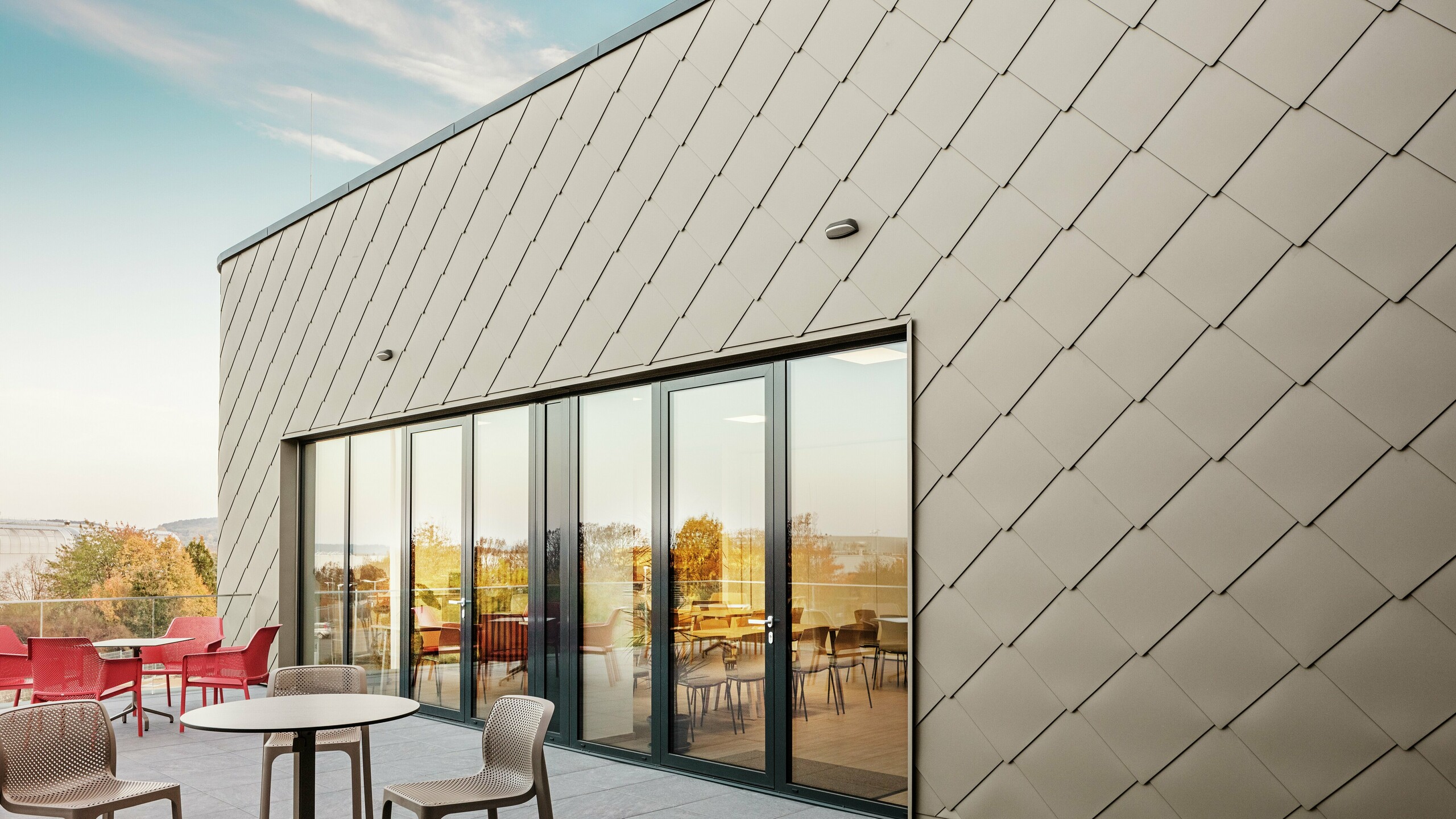 Die Fassade des neuen PREFA Standortes in Törökbálint besticht durch die eleganten PREFA Wandrauten 44 × 44 in P.10 Prefabronze. Die präzise verlegte Aluminiumfassade sorgt für ein modernes, dynamisches Erscheinungsbild und bietet gleichzeitig maximalen Schutz vor Witterungseinflüssen. Die großzügige Glasfront verbindet Innen- und Außenbereich und schafft auf der großzügigen Terrasse eine helle und einladende Atmosphäre.