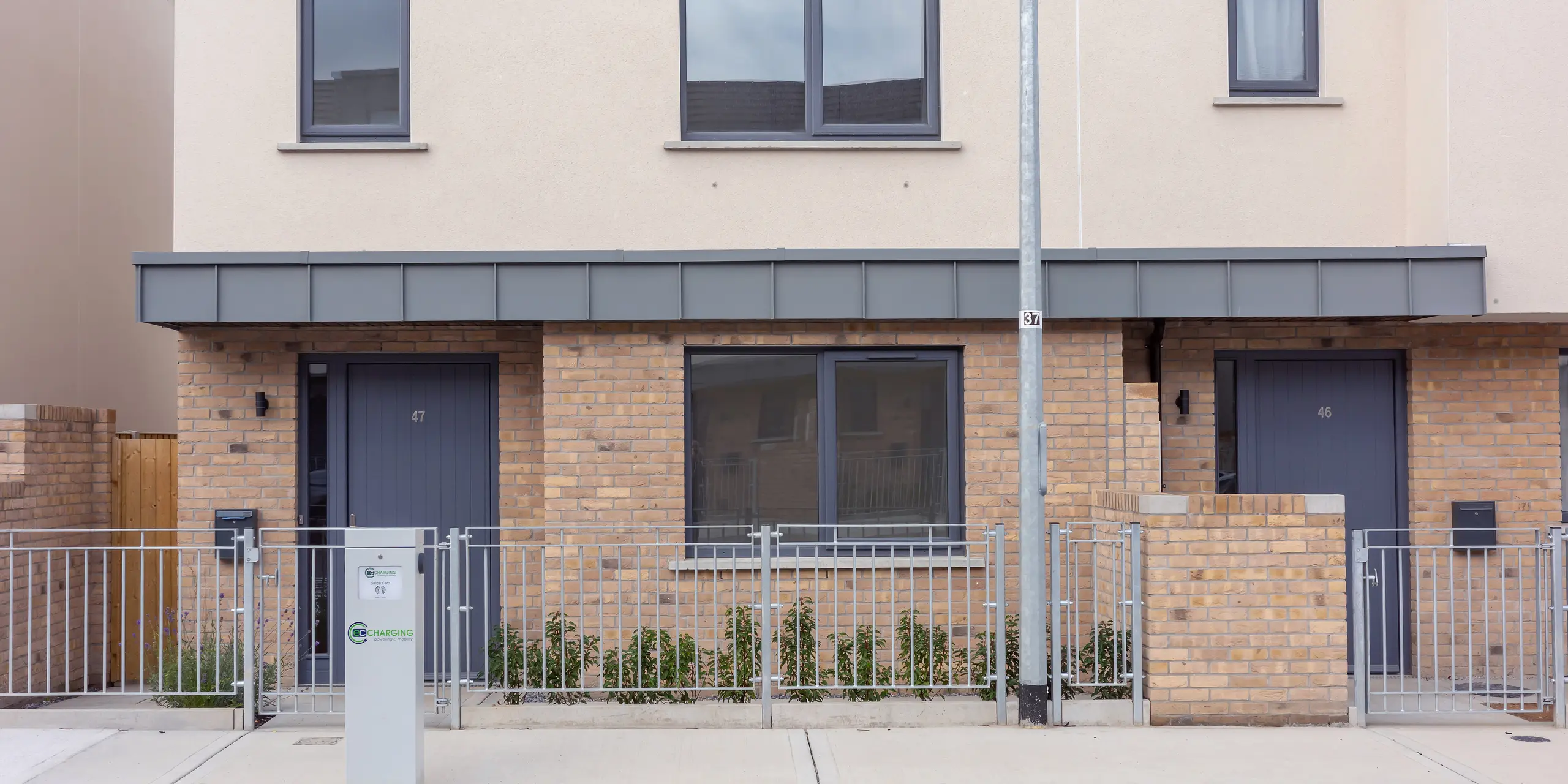 Das Bild zeigt den Eingangsbereich einer modernen Reihenhausanlage in Churchfields, geprägt von klaren Linien und harmonischen Farbflächen. Besonders ins Auge fällt die elegante Überdachung über den Haustüren, die mit PREFALZ in P.10 Hellgrau verkleidet ist. Die robuste Aluminiumoberfläche setzt einen modernen Akzent über dem warmen Backstein und ist nahezu wartungsfrei – ideal für große Wohnprojekte. Die einheitliche Fassadengestaltung und der gepflegte Vorgarten verleihen der Szene eine ruhige Ausstrahlung.