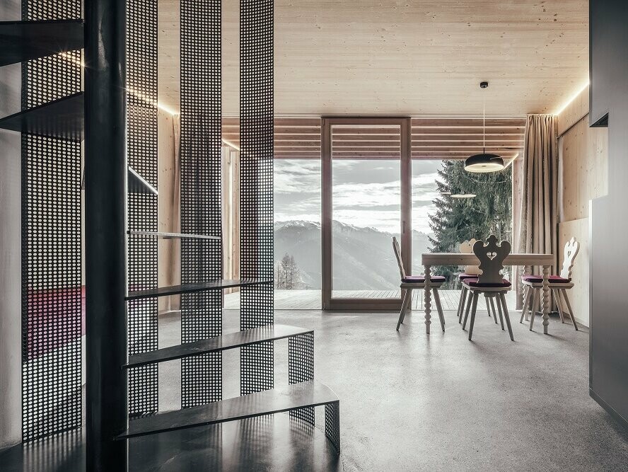 Photo de l’intérieur. Sont visibles : l’escalier en colimaçon, le sol en béton apparent, les meubles de menuisier au style rustique. Vue sur les Dolomites au-dessus du grand balcon.