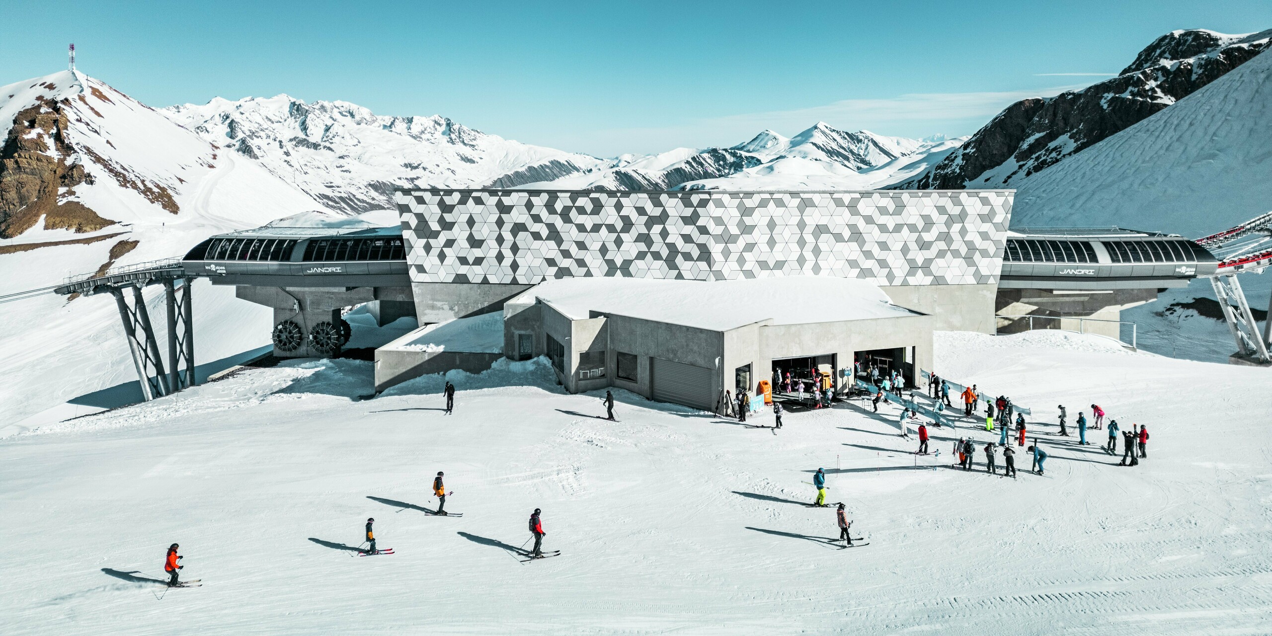 Die futuristische Seilbahnstation in Les Deux Alpes, umgeben von einer atemberaubenden Berglandschaft, präsentiert sich mit einer auffälligen Fassade aus PREFABOND Aluminium Verbundplatten. Das markante, geometrische Rautenmuster in Weiß und Grau fügt sich perfekt in die schneebedeckte Umgebung ein. Die langlebigen und wetterbeständigen Materialien gewährleisten eine ästhetisch ansprechende und funktionale Lösung für die extremen Bedingungen im Hochgebirge. Vor der Mittelstation bereiten sich zahlreiche Skifahrer und Snowboarder auf die Abfahrt vor.