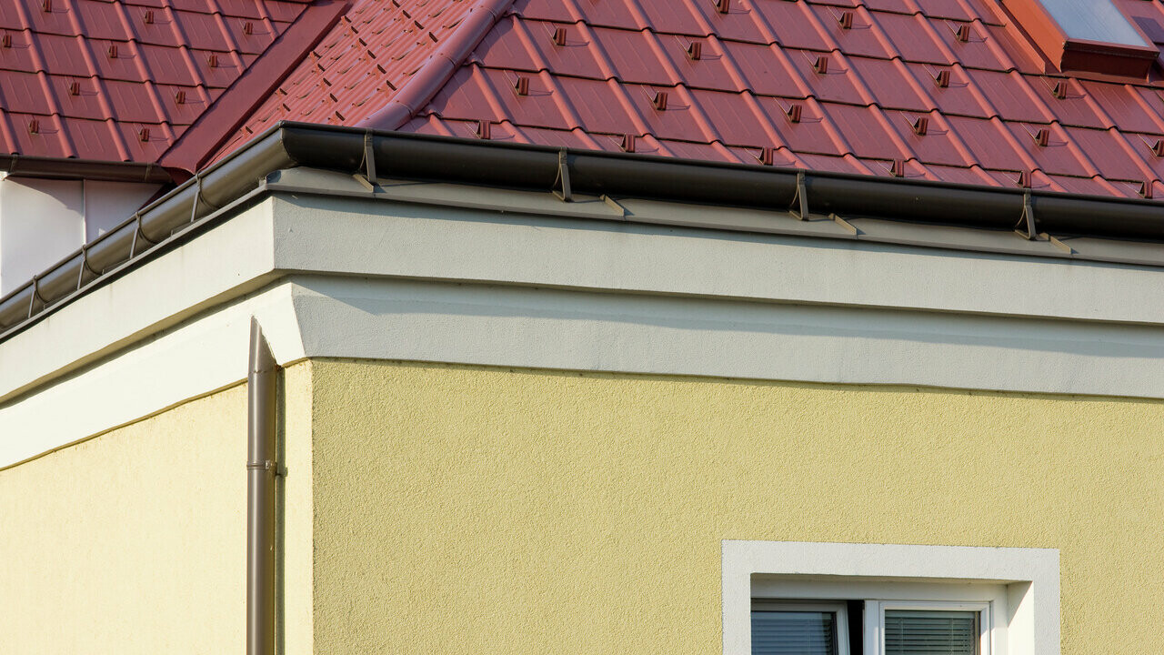 Angle de maison avec façade en crépi jaune et gouttière havraise (gouttière sur toiture) PREFA en brun ; le toit est en tuiles PREFA rouge