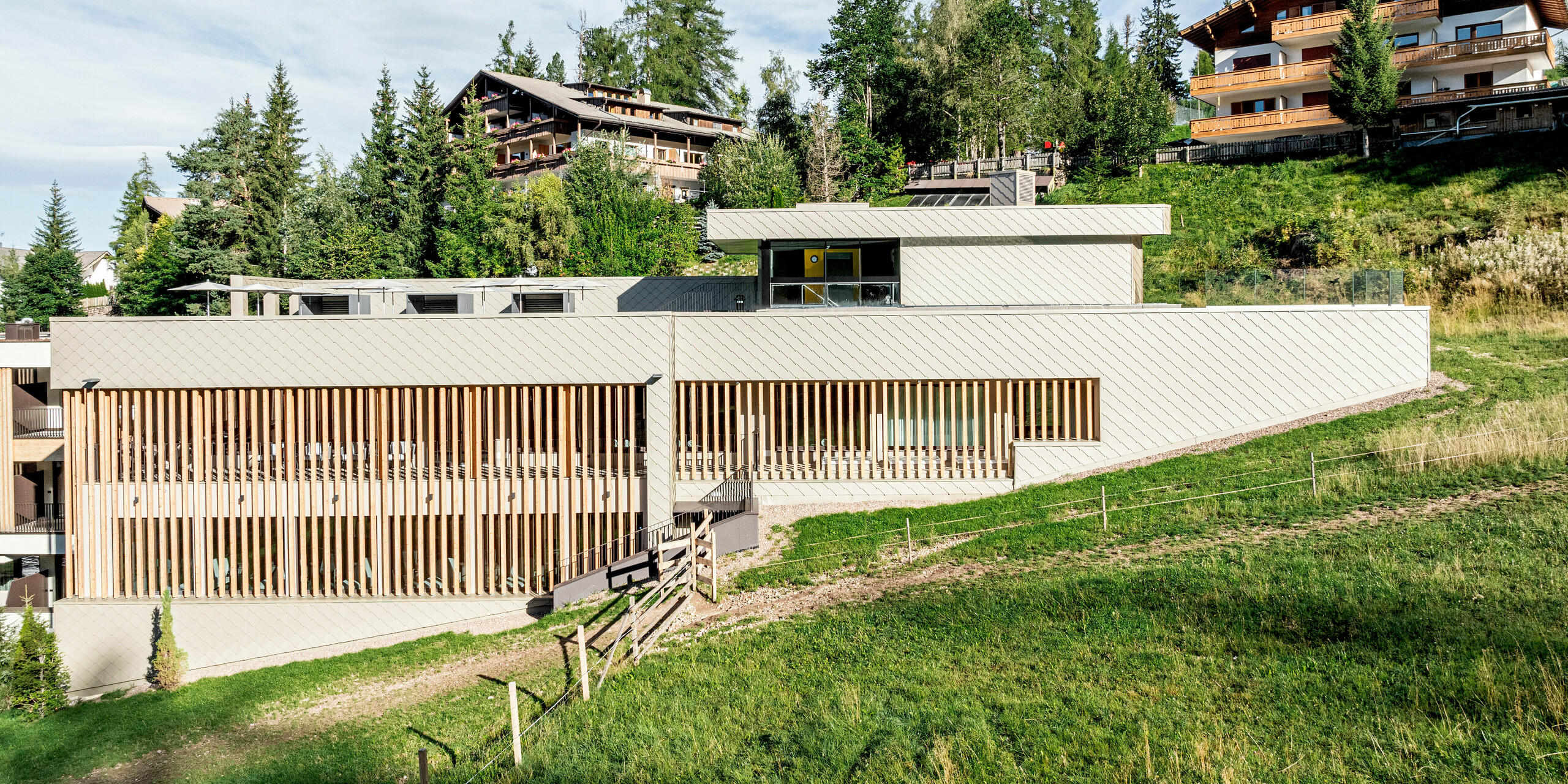 Seitenansicht des Sporthotels Obereggen in Südtirol, eingebettet in eine sanfte alpine Hanglage mit Wiesen, Bäumen und umliegender Bergbebauung. Der langgestreckte Baukörper wird durch die PREFA Aluminiumfassade mit Wandraute 29 × 29 in der Farbe Bronze geprägt, die der Architektur eine klare Linienführung und lebendige Struktur verleiht. Der warme Bronzeton kontrastiert harmonisch mit Holzlamellen und Natur. Die langlebigen, witterungsbeständigen und wartungsarmen PREFA Aluminiumprodukte sind ideal für anspruchsvolle Architektur im alpinen Raum.