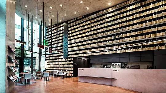 [Translate to Italiano:] Das Bild zeigt das moderne und stilvolle Innere der Bibliothek in Olmütz, das durch seine beeindruckende Bücherwand dominiert wird. Die bis zur Decke reichenden Regale erzeugen eine dramatische Wirkung, während der schlichte, aber elegante Empfangstresen aus feinstem Beton und Holz eine angenehme Atmosphäre schafft. Besonders hervorzuheben sind die klaren Linien und die präzise Beleuchtung, die das Gesamtbild harmonisch ergänzen. Die Verwendung von PREFABOND Aluminium Verbundplatten und anderen hochwertigen Materialien an der Außenfassade sorgt für eine langlebige und pflegeleichte Gestaltung. Diese Architektur verbindet Funktionalität und ästhetischen Anspruch auf eindrucksvolle Weise.