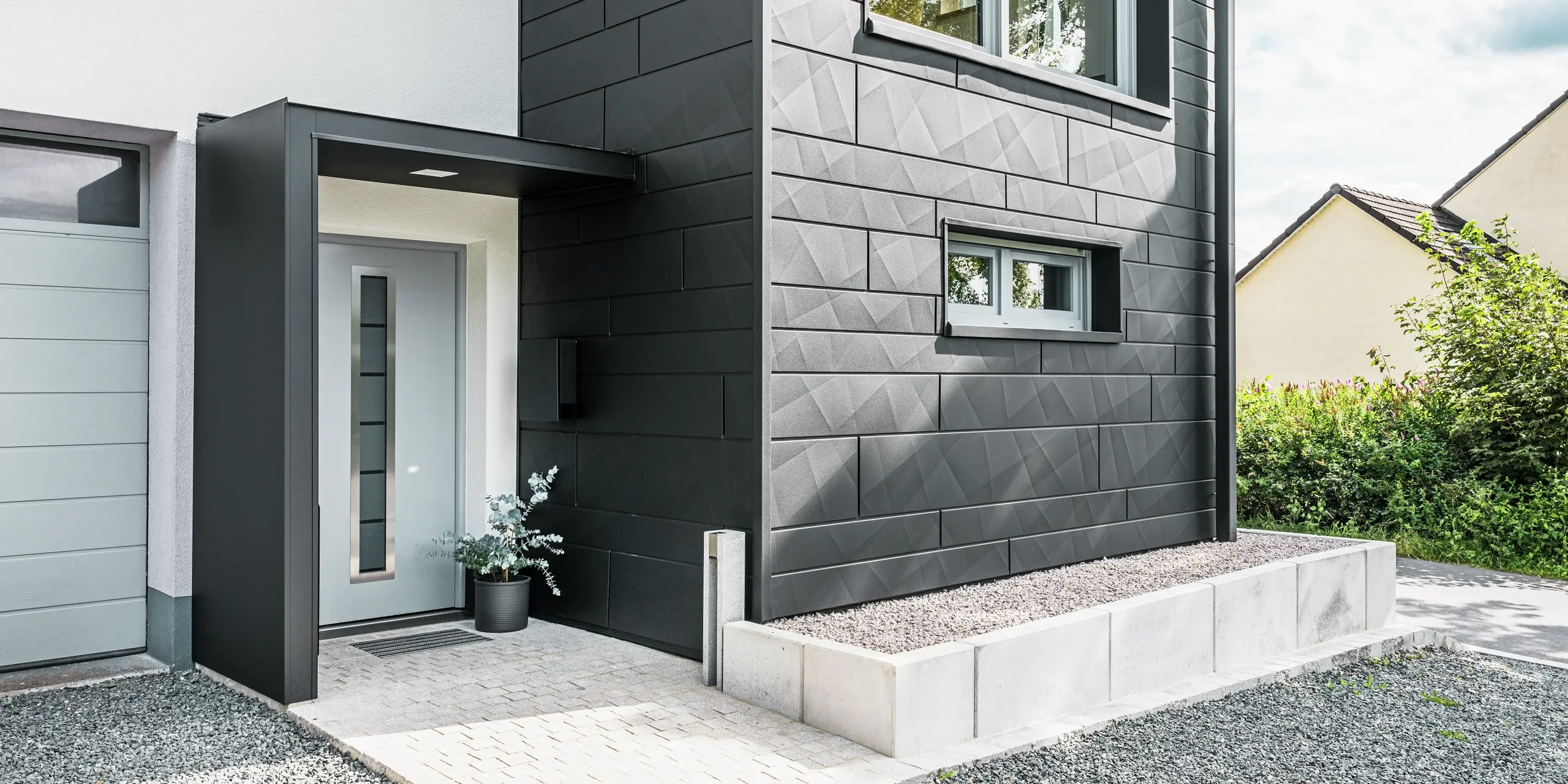 Die elegante PREFA Siding.X Fassade in P.10 Schwarz dominiert den Eingangsbereich des modernisierten Einfamilienhauses in Teuschnitz. Die hochwertige Blechfassade verleiht dem Gebäude einen modernen und zeitlosen Charakter. Die klar strukturierten Fassadenpaneele schaffen ein markantes, geometrisches Design, während die schwarzen Aluminium Verbundplatten die Eingangstür und die Fenster umrahmen. Das PREFA Quadratrohr fügt sich nahtlos in das anspruchsvolle Design ein und unterstreicht die funktionale und ästhetische Wirkung der Sanierung. Der Weg zum Haus ist mit Pflastersteinen und Kies versehen.