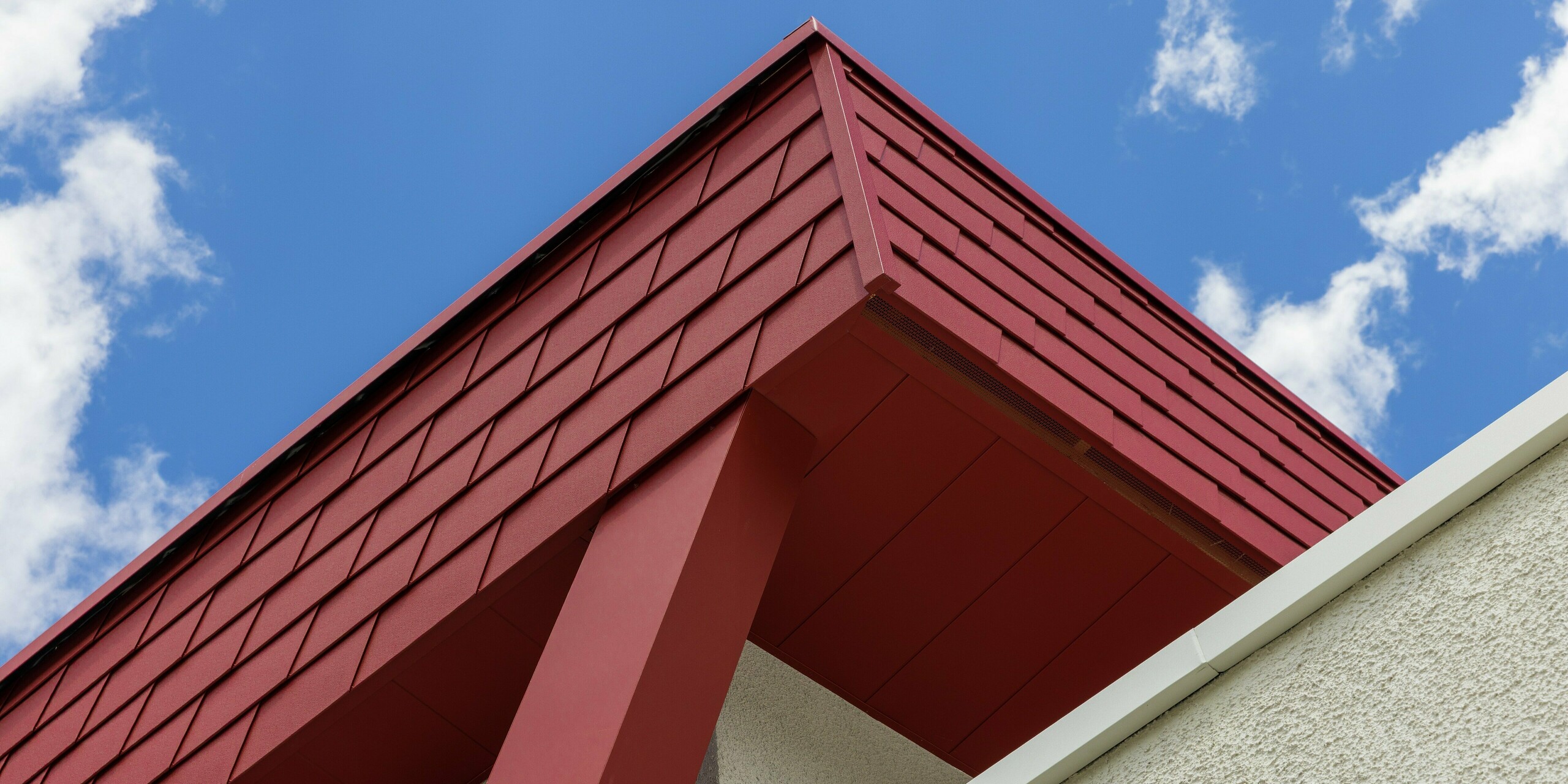 Détail de la façade du Summit Mountain Club à Ratschings, revêtue de robustes bardeaux PREFA de couleur Rouge Oxyde P.10 et d'une poutre de soutien rouge du plus bel effet. La façade métallique PREFA de haute qualité est non seulement esthétique, mais offre également une protection durable contre les rigueurs du climat alpin.