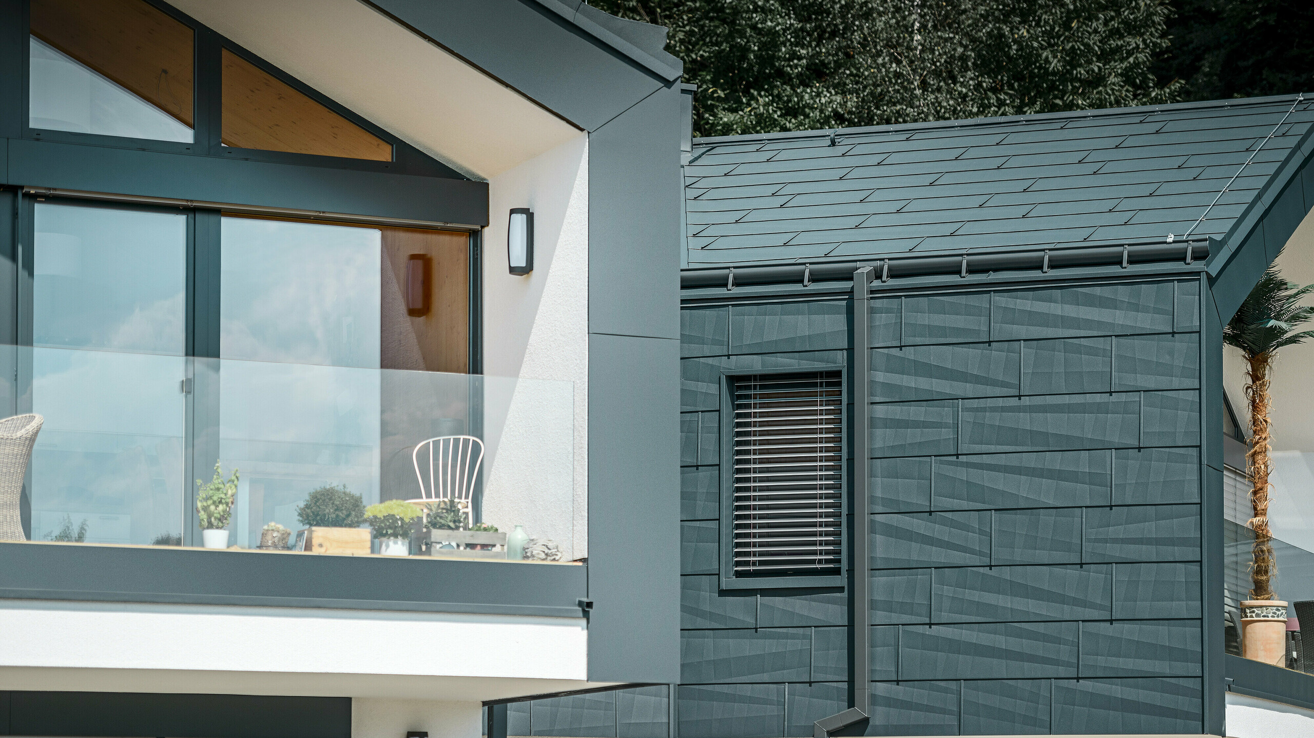 La photo montre une maison jumelée moderne avec une façade élégante en aluminium. La façade est peinte dans un ton anthracite foncé et présente une finition structurée grâce au panneau de toiture et de façade PREFA FX.12. Une gouttière havraise et un tuyau de descente carré PREFA ont été installés sur le toit. Un balcon avec une balustrade en verre est visible à l'étage supérieur, tandis que l'étage inférieur est doté de fenêtres avec des stores à lamelles. L'architecture est claire et minimaliste, et des arbres sont visibles en arrière-plan, créant une atmosphère naturelle.
