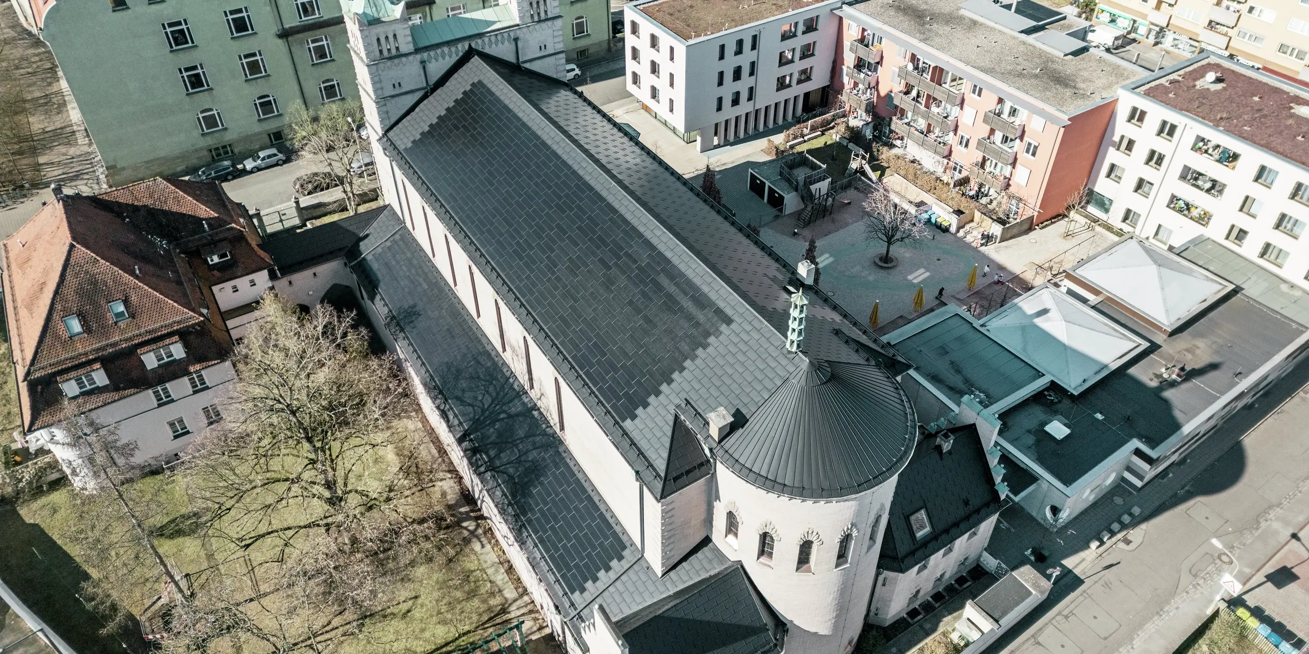 Vogelperspektive auf die Rückseite der St. Karl Borromäus Kirche in Nürnberg. Die schwarzen PREFA Solardachplatten decken das markante, runde Satteldach über dem Hauptteil der Kirche als auch das Dach des Nebengebäudes. Die Aluminiumdachplatten mit integrierten PV-Modulen fügen sich perfekt in das historische Erscheinungsbild der Kirche ein. Der Blick auf das moderne Umfeld mit angrenzenden Wohngebäuden und dem grünen Hofbereich verdeutlicht die gelungene Verbindung von Denkmalschutz, moderner Architektur und nachhaltiger Energiegewinnung. Eine ideale Lösung für den Denkmalschutz im urbanen Raum.