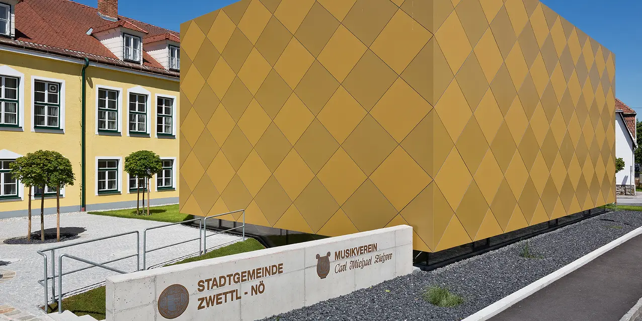 Eingangsbereich zum Musikverein Zwettl. Vor dem Gebäude ist die Funktion des Objekts in Stein gemeißelt. Das quaderförmige Gebäude besticht durch die markante Optik mit PREFA Aluminium Verbundplatten in zwei unterschiedlichen Goldtönen. Die Aluminiumplatten sind sorgfältig in Form von Karos auf der Fassade montiert worden. 
