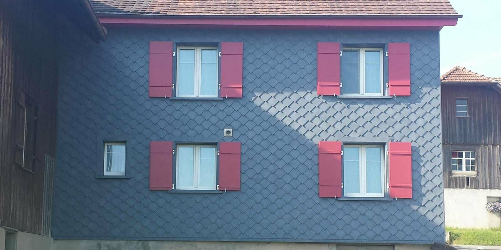 Rénovation de façade à l’aide de losanges de façade PREFA, avec quatre fenêtres dotées de volets rouges