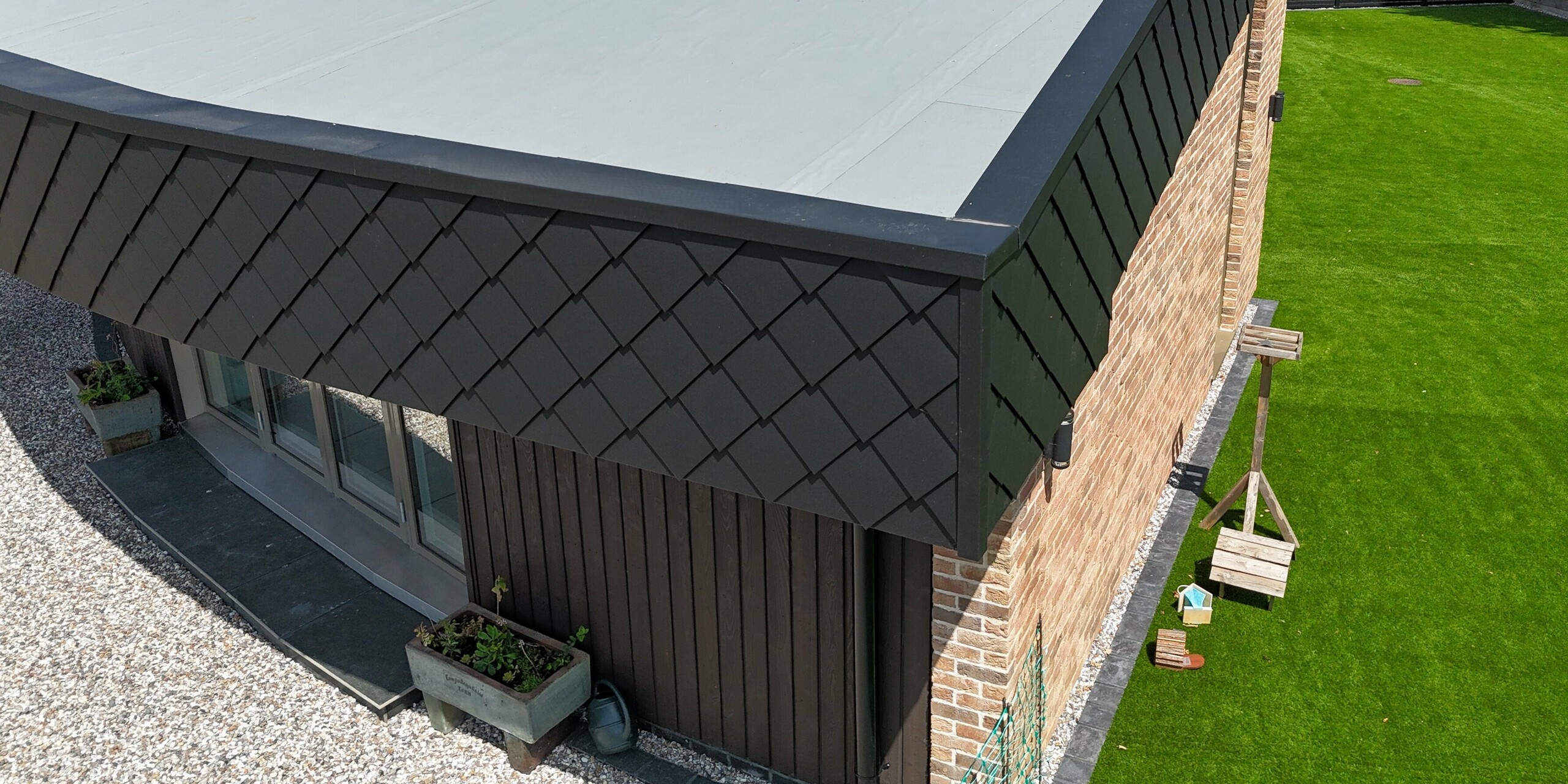 Vue détaillée d'une maison écologique moderne à la façade incurvée remarquable, réalisée grâce à l'association de briques de parement chaleureuses et de l'élégant bardage PREFA en losanges 20 x 20 e P.10 anthracite. Les éléments en aluminium créent un contraste saisissant et confèrent à la maison une allure dynamique et haut de gamme. Grâce à la robustesse des produits PREFA, la façade est extrêmement durable, résistante aux intempéries et facile d'entretien – idéale pour une architecture durable. La pureté des lignes et la précision de la réalisation sont clairement visibles, soulignant parfaitement l'harmonie entre design et fonctionnalité.