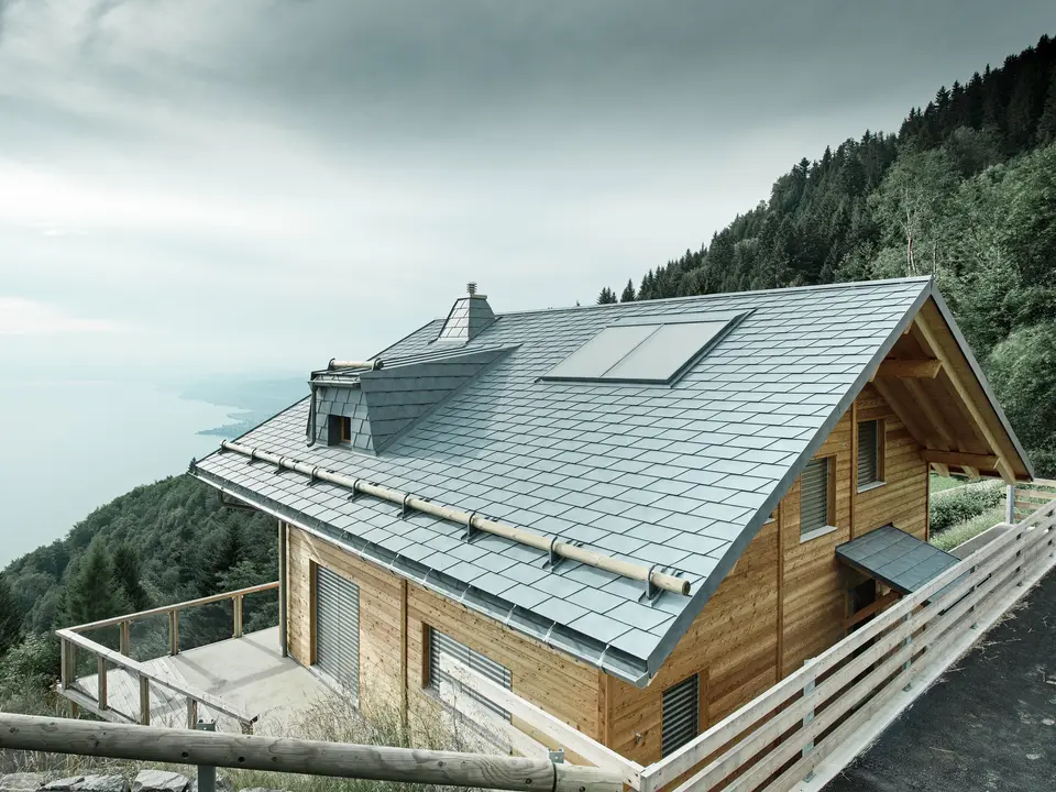 Dachansicht einer Holz-Lodge am Genfersee mit PREFA Dachschindeln in P.10 Steingrau. Die präzise verarbeitete Dachgaube und der Kamin in Trapezform setzen markante Akzente, während das robuste Dach Wind und Wetter trotzt. Das elegante Design aus Aluminium in Kombination mit Holz sorgt für eine perfekte Harmonie zwischen Funktion und Ästhetik. Im Hintergrund ist der in mystischen Nebel gehüllte Genfersee zu erkennen.