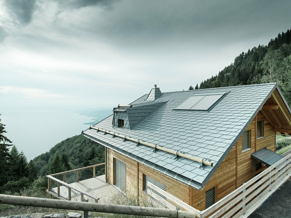 Dachansicht einer Holz-Lodge am Genfersee mit PREFA Dachschindeln in P.10 Steingrau. Die präzise verarbeitete Dachgaube und der Kamin in Trapezform setzen markante Akzente, während das robuste Dach Wind und Wetter trotzt. Das elegante Design aus Aluminium in Kombination mit Holz sorgt für eine perfekte Harmonie zwischen Funktion und Ästhetik. Im Hintergrund ist der in mystischen Nebel gehüllte Genfersee zu erkennen.
