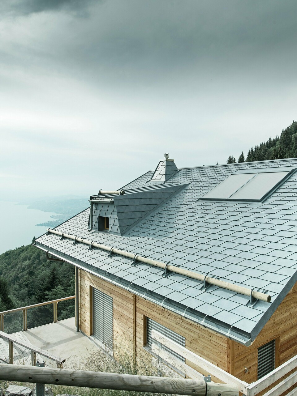 Dachansicht einer Holz-Lodge am Genfersee mit PREFA Dachschindeln in P.10 Steingrau. Die präzise verarbeitete Dachgaube und der Kamin in Trapezform setzen markante Akzente, während das robuste Dach Wind und Wetter trotzt. Das elegante Design aus Aluminium in Kombination mit Holz sorgt für eine perfekte Harmonie zwischen Funktion und Ästhetik. Im Hintergrund ist der in mystischen Nebel gehüllte Genfersee zu erkennen.