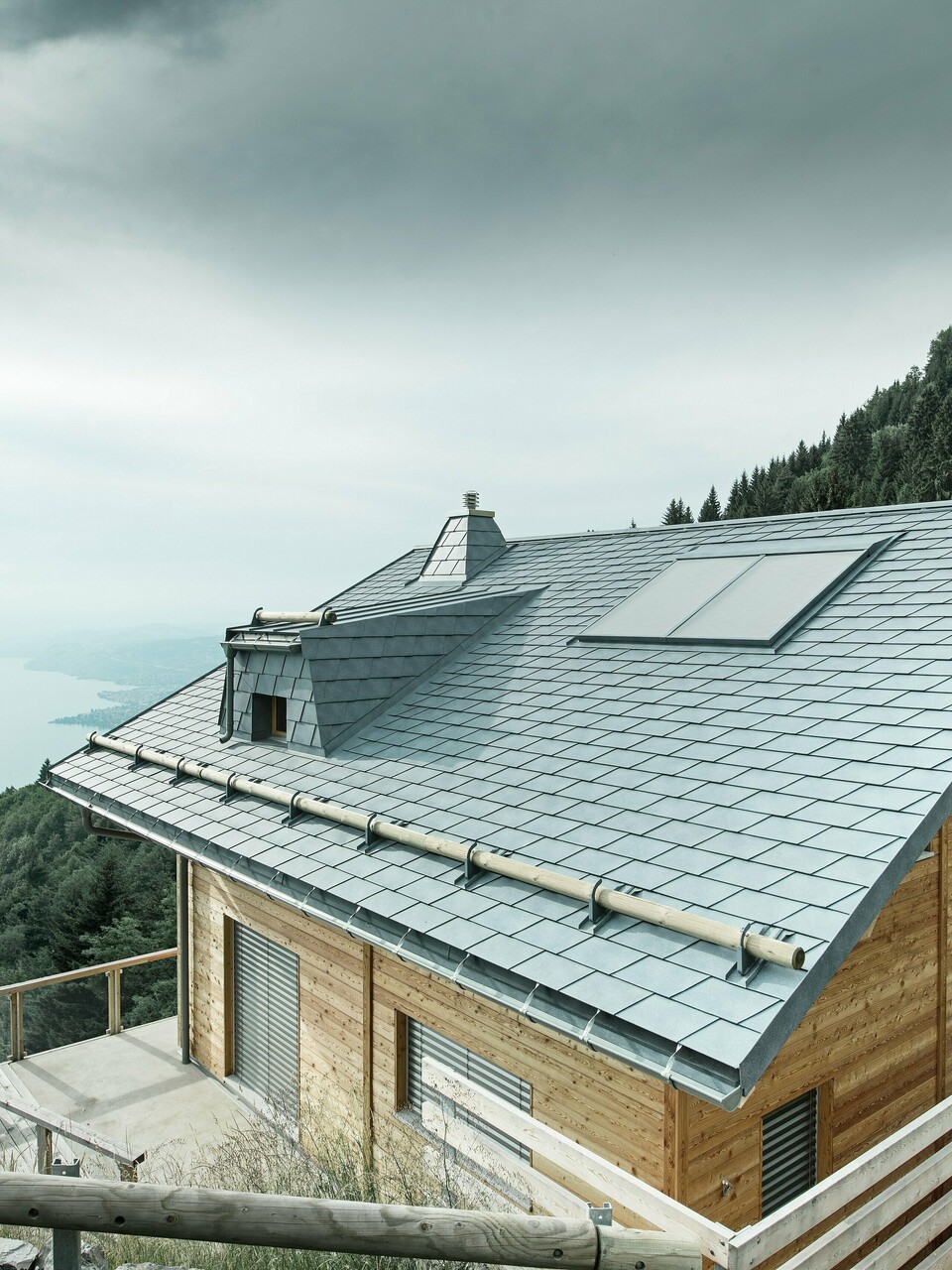 Dachansicht einer Holz-Lodge am Genfersee mit PREFA Dachschindeln in P.10 Steingrau. Die präzise verarbeitete Dachgaube und der Kamin in Trapezform setzen markante Akzente, während das robuste Dach Wind und Wetter trotzt. Das elegante Design aus Aluminium in Kombination mit Holz sorgt für eine perfekte Harmonie zwischen Funktion und Ästhetik. Im Hintergrund ist der in mystischen Nebel gehüllte Genfersee zu erkennen.