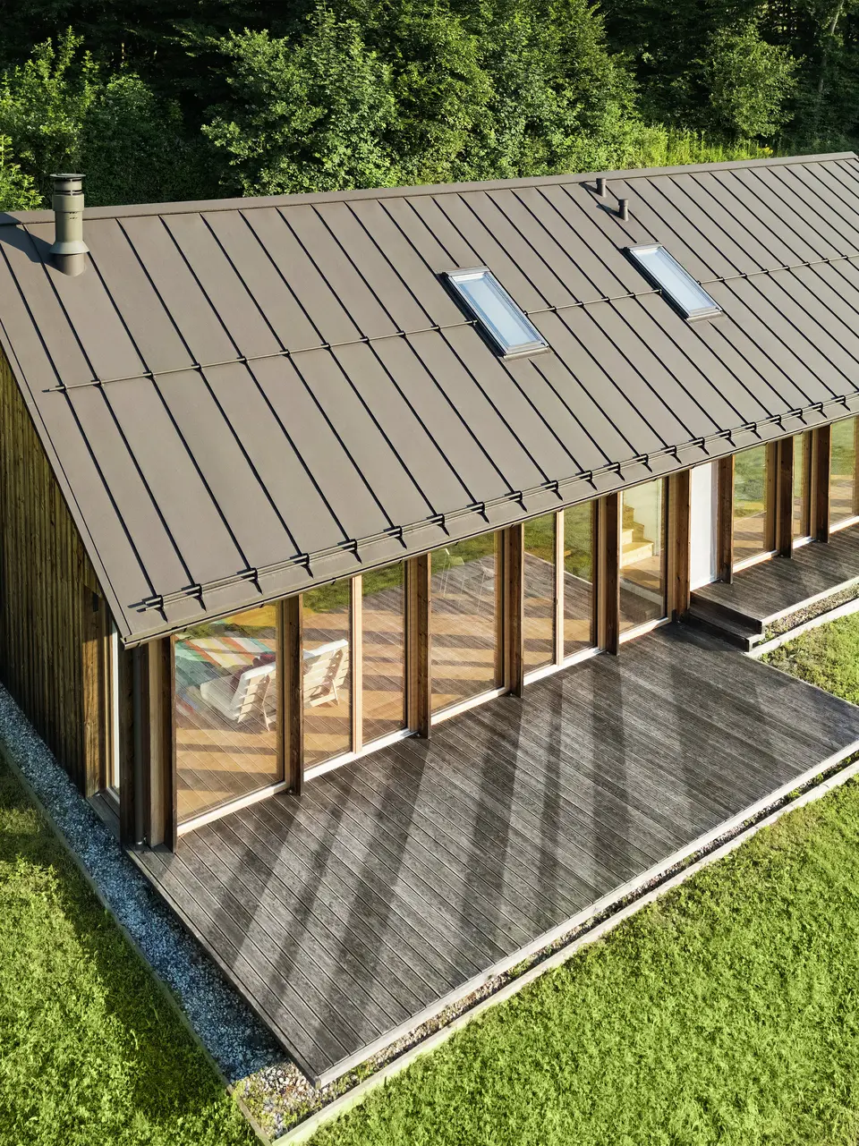 Moderna casa in legno con generosa facciata in vetro e resistente tetto in alluminio PREFALZ in P.10 marrone - ideale per siti in pendenza come Vinje. La copertura del tetto di alta qualità garantisce la massima resistenza agli agenti atmosferici, una lunga durata e un aspetto elegante. L'installazione precisa delle giunture verticali e i lucernari integrati creano un concetto abitativo sostenibile e inondato di luce. La vista a volo d'uccello mostra la precisa lavorazione delle aggraffature in alluminio.