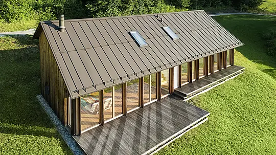 Moderna casa in legno con generosa facciata in vetro e resistente tetto in alluminio PREFALZ in P.10 marrone - ideale per siti in pendenza come Vinje. La copertura del tetto di alta qualità garantisce la massima resistenza agli agenti atmosferici, una lunga durata e un aspetto elegante. L'installazione precisa delle giunture verticali e i lucernari integrati creano un concetto abitativo sostenibile e inondato di luce. La vista a volo d'uccello mostra la precisa lavorazione delle aggraffature in alluminio.