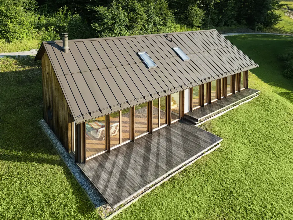 Moderna casa in legno con generosa facciata in vetro e resistente tetto in alluminio PREFALZ in P.10 marrone - ideale per siti in pendenza come Vinje. La copertura del tetto di alta qualità garantisce la massima resistenza agli agenti atmosferici, una lunga durata e un aspetto elegante. L'installazione precisa delle giunture verticali e i lucernari integrati creano un concetto abitativo sostenibile e inondato di luce. La vista a volo d'uccello mostra la precisa lavorazione delle aggraffature in alluminio.
