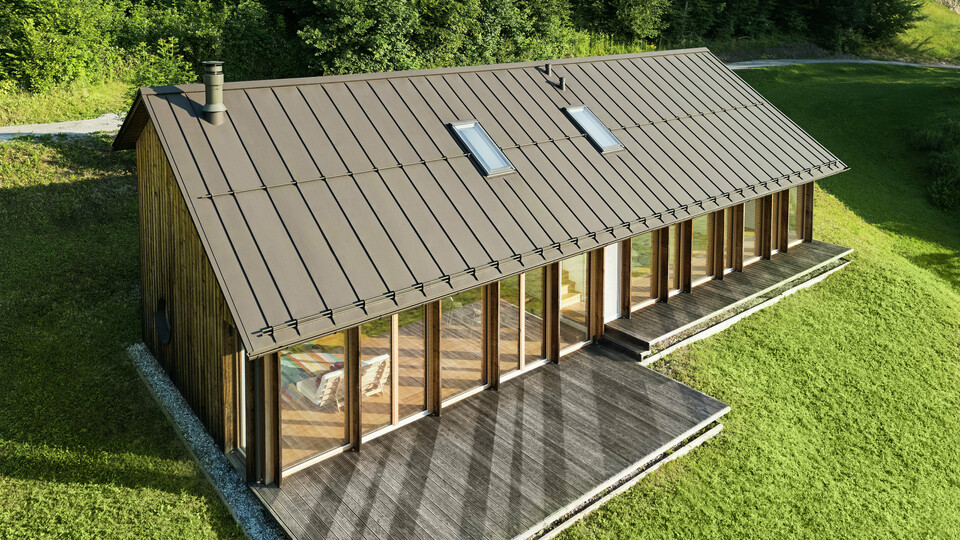Maison en bois moderne avec une grande façade vitrée et un toit en aluminium PREFALZ résistant de couleur P.10 brun – idéale pour les terrains en pente comme à Vinje. La couverture de toit de haute qualité offre une résistance maximale aux intempéries, une longue durée de vie et une esthétique élégante. Grâce à la pose précise des joints debout et à l’intégration de fenêtres de toit, un concept d’habitat durable et baigné de lumière voit le jour. La vue en plongée met en évidence la finition soignée des joints debout en aluminium.