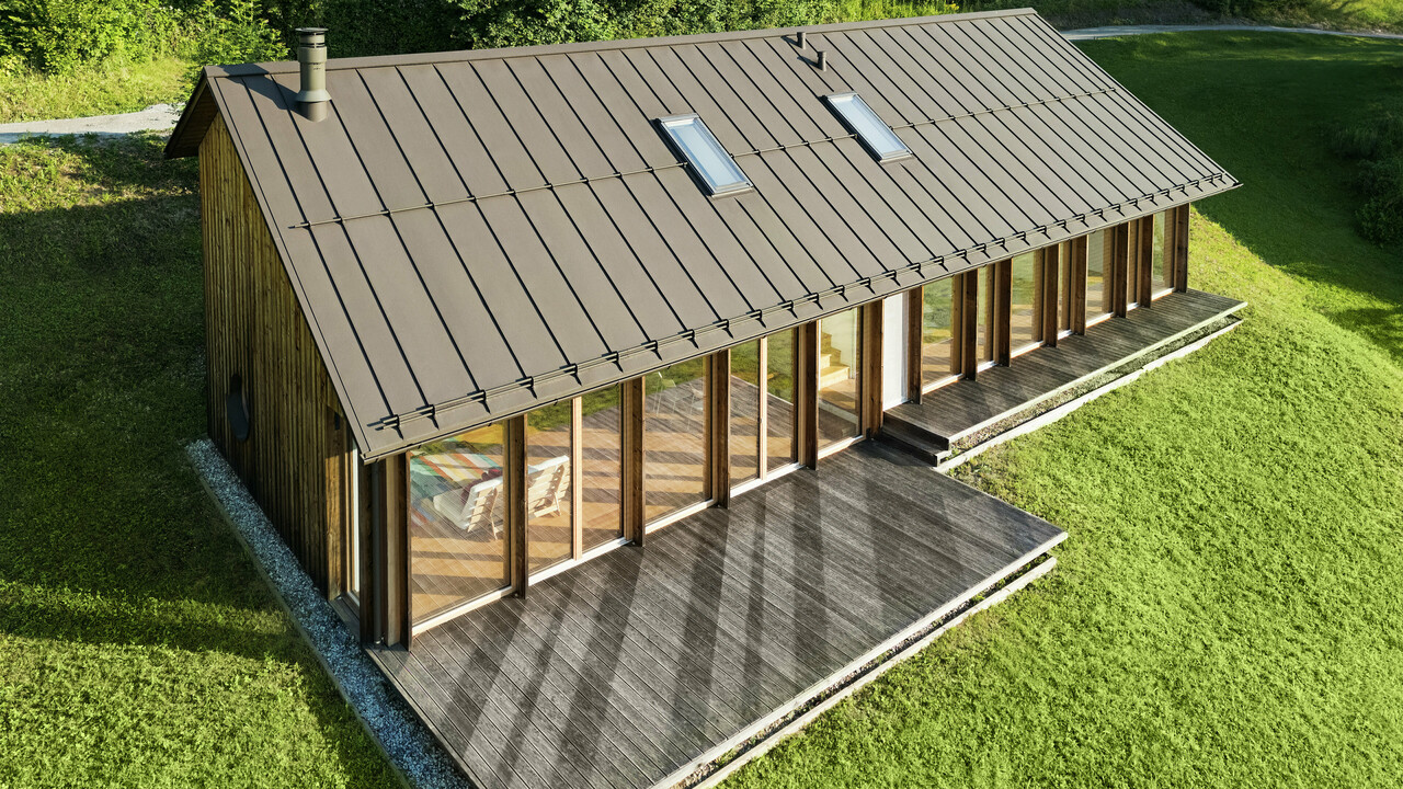 Maison en bois moderne avec une grande façade vitrée et un toit en aluminium PREFALZ résistant de couleur P.10 brun – idéale pour les terrains en pente comme à Vinje. La couverture de toit de haute qualité offre une résistance maximale aux intempéries, une longue durée de vie et une esthétique élégante. Grâce à la pose précise des joints debout et à l’intégration de fenêtres de toit, un concept d’habitat durable et baigné de lumière voit le jour. La vue en plongée met en évidence la finition soignée des joints debout en aluminium.