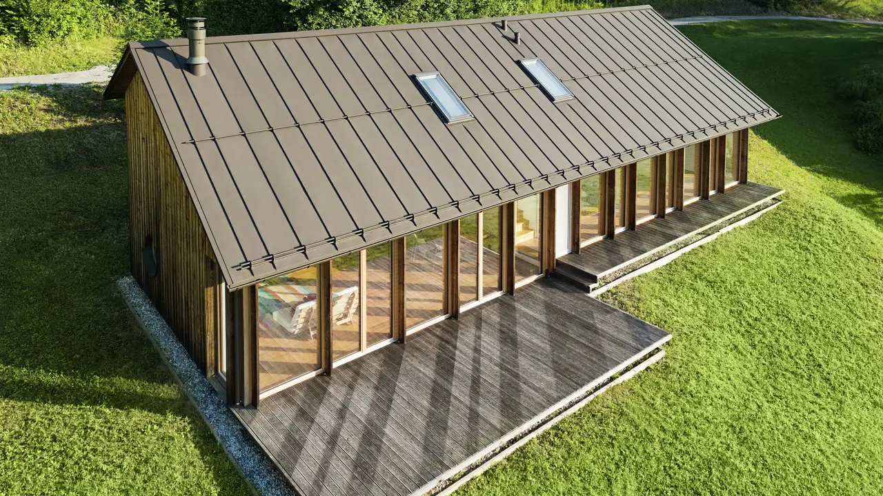 Moderna casa in legno con generosa facciata in vetro e resistente tetto in alluminio PREFALZ in P.10 marrone - ideale per siti in pendenza come Vinje. La copertura del tetto di alta qualità garantisce la massima resistenza agli agenti atmosferici, una lunga durata e un aspetto elegante. L'installazione precisa delle giunture verticali e i lucernari integrati creano un concetto abitativo sostenibile e inondato di luce. La vista a volo d'uccello mostra la precisa lavorazione delle aggraffature in alluminio.