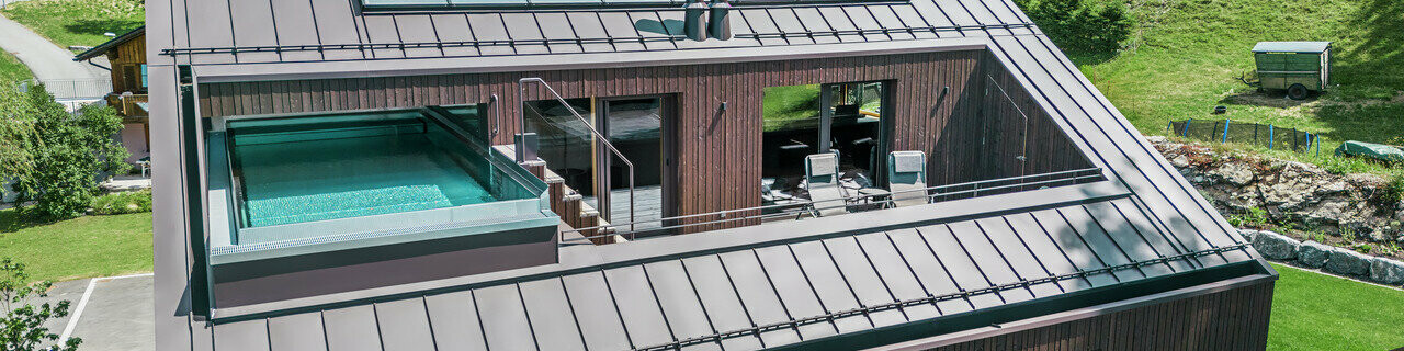 Vue aérienne d'une maison moderne avec un toit en Prefalz PREFA brun noisette. Un toit-terrasse avec piscine offre un espace de détente exclusif avec vue sur le paysage vallonné et verdoyant.