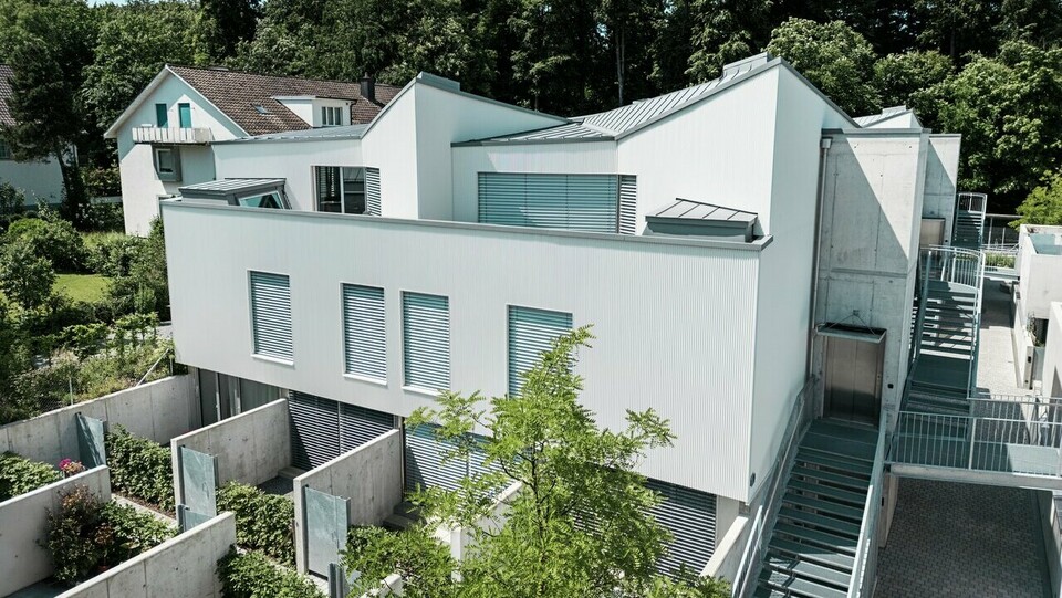 Immeuble collectif à l'architecture moderne, doté d'une façade verticale à profil triangle gris quartz et d'une toiture PREFALZ dans une couleur assortie. Les lignes épurées du bâtiment s'harmonisent avec les espaces extérieurs intégrés, les éléments en béton et les espaces verts structurés au rez-de-chaussée. À l'arrière-plan, une végétation dense et une maison traditionnelle soulignent le contraste avec l'architecture moderne.