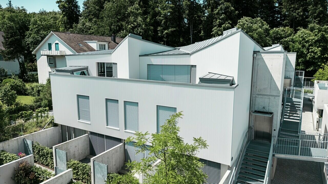Mehrfamilienhaus mit moderner Architektur, ausgestattet mit einer vertikalen Fassade aus Zackenprofil in Patinagrau und einem Dach aus PREFALZ in passender Farbgebung. Die klaren Linien des Gebäudes harmonieren mit den integrierten Außenbereichen, Betonelementen und den strukturierten Gartenflächen im Erdgeschoss. Im Hintergrund dichter Baumbestand und ein traditionelles Wohnhaus, die den Kontrast zur modernen Bauweise unterstreichen.