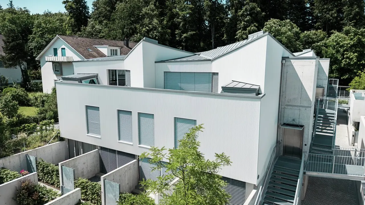Mehrfamilienhaus mit moderner Architektur, ausgestattet mit einer vertikalen Fassade aus Zackenprofil in Patinagrau und einem Dach aus PREFALZ in passender Farbgebung. Die klaren Linien des Gebäudes harmonieren mit den integrierten Außenbereichen, Betonelementen und den strukturierten Gartenflächen im Erdgeschoss. Im Hintergrund dichter Baumbestand und ein traditionelles Wohnhaus, die den Kontrast zur modernen Bauweise unterstreichen.