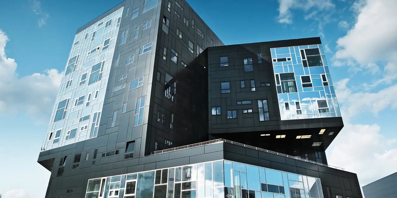 Modernes Universitätsgebäude mit markanter, geometrischer Architektur und überhängenden Baukörpern. Die Fassade besteht aus dunklen und hellen Aluminium-Verbundplatten in unterschiedlichen Grautönen mit unregelmäßig angeordneten Fenstern. Das Gebäude steht unter blauem Himmel mit vereinzelten Wolken und spiegelt das Licht in verschiedenen Facetten wider.