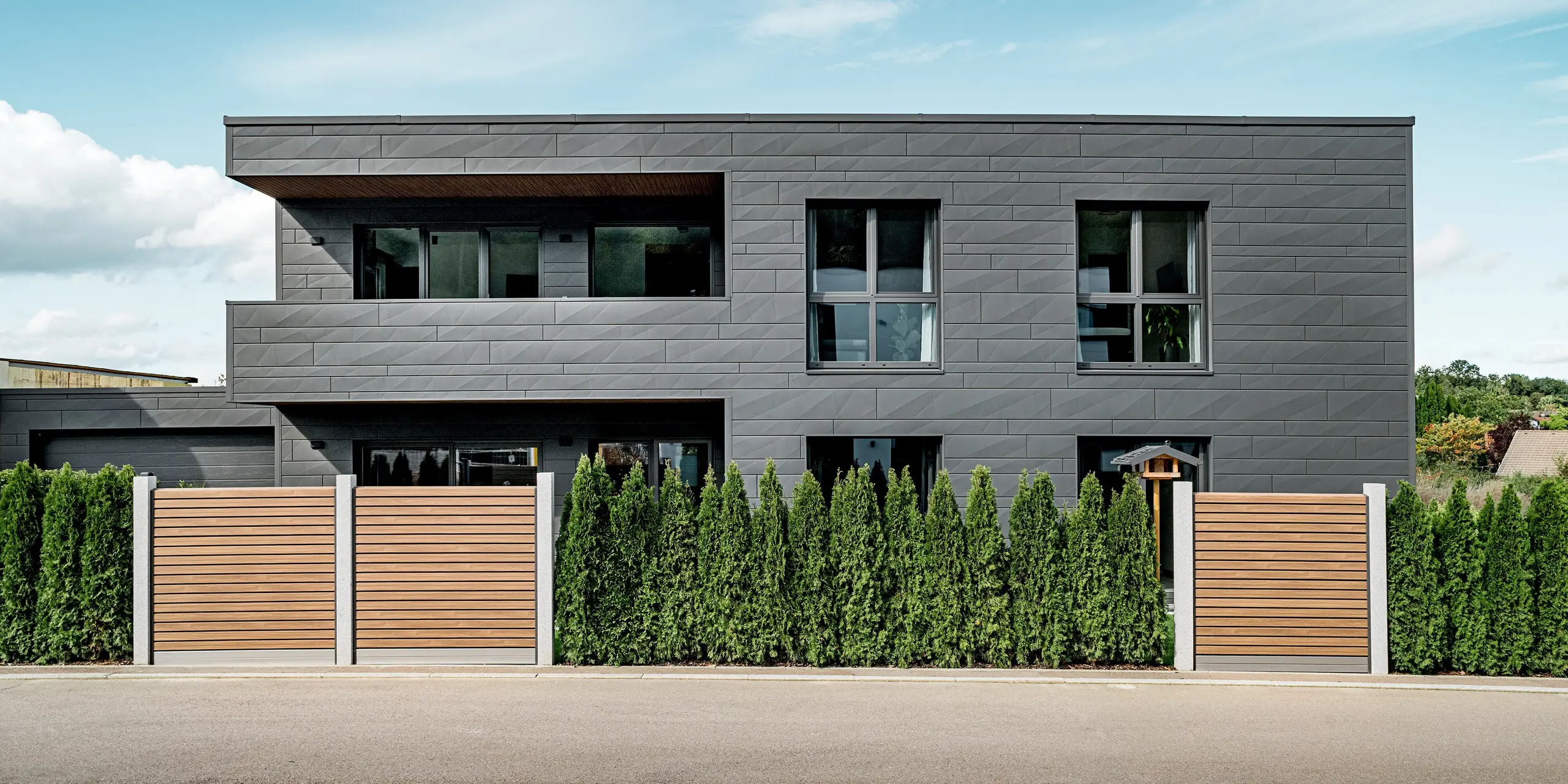 Vue de rue d'une maison individuelle moderne à Heidenheim, dont la façade est entièrement revêtue de panneaux d'aluminium Siding.X PREFA, coloris P.10 gris sombre. Les panneaux aux lignes nettes, de largeurs variables et aux arêtes marquées, créent un effet géométrique et dynamique. Au premier plan, une haie de thuyas dense longe une clôture en bois à lattes verticales, offrant un contraste chaleureux avec la froideur de l'architecture métallique. De larges fenêtres reflètent le ciel et les environs. La surface en aluminium PREFA, résistante et durable, garantit une protection optimale et un style résolument moderne.