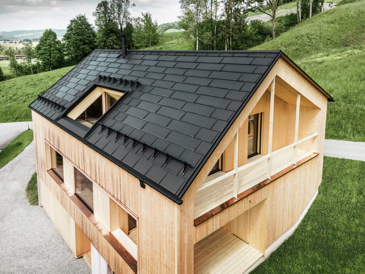 Vista dall'alto di una casa ecologica in legno a Egg, immersa in prati verdi, con un tetto PREFA Solar scuro e resistente. Le tegole nere del tetto in alluminio R.16 si armonizzano con i moduli fotovoltaici, integrati in modo efficiente nel tetto della casa indipendente per generare energia sostenibile. L'edificio, con la sua caratteristica facciata in legno e l'architettura moderna, emana naturalezza e design orientato al futuro, rispettando al contempo il paesaggio con la sua avanzata tecnologia solare.