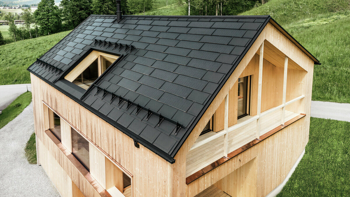 Vue aérienne d'une maison en bois écologique à Egg, nichée dans des prairies verdoyantes, avec un toit solaire PREFA sombre et durable. Les R.16 PREFA en aluminium noirs s'harmonisent avec les modules solaires, qui sont efficacement intégrés dans le toit de la maison unifamiliale pour générer une énergie durable. Le bâtiment, avec sa façade en bois distinctive et son architecture moderne, respire le naturel et un design avant-gardiste tout en respectant le paysage grâce à sa technologie solaire avancée.