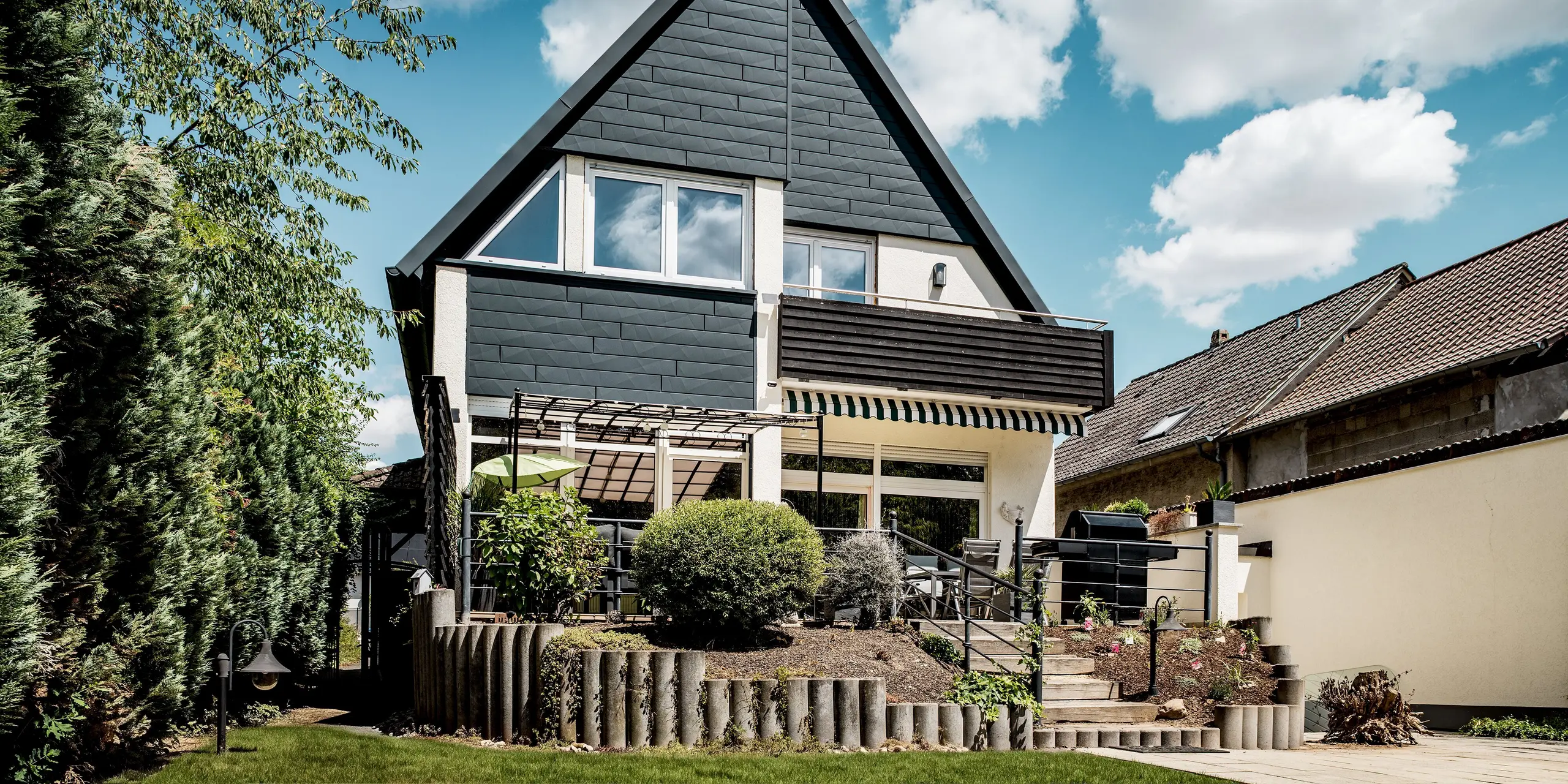 Modernes Einfamilienhaus in Hargesheim mit markanter Giebelfassade aus PREFA Siding.X in P.10 Anthrazit. Die strukturierte Aluminiumverkleidung setzt spannende Akzente und bildet einen eleganten Kontrast zur hellen Putzfassade und großzügigen Fensterfront. Die leichte, langlebige und witterungsbeständige Fassadenlösung unterstreicht den modernen Charakter des sanierten Wohnhauses und sorgt für dauerhaften Schutz.