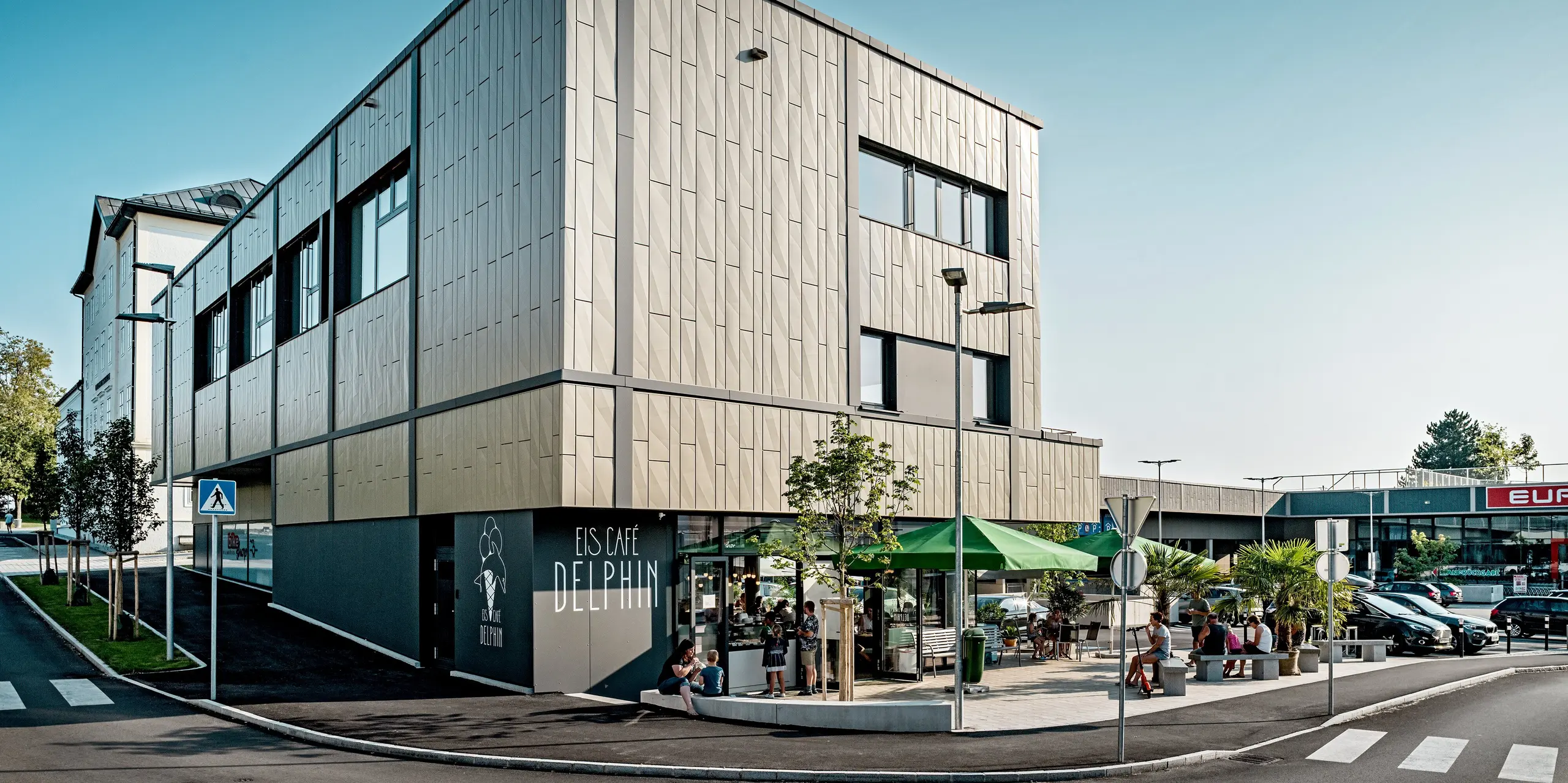L'image présente l'angle moderne du centre commercial de Rohrbach, avec son élégante façade en aluminium signée PREFA. Cette façade est composée de panneaux Siding.X bronze et anthracite. Leur texture fine et leur design géométrique créent un effet saisissant. Devant le bâtiment se trouve le glacier Delphin, doté d'une terrasse accueillante et d'espaces verts qui contribuent à une atmosphère agréable. La façade s'intègre harmonieusement à son environnement, alliant fonctionnalité et design contemporain.