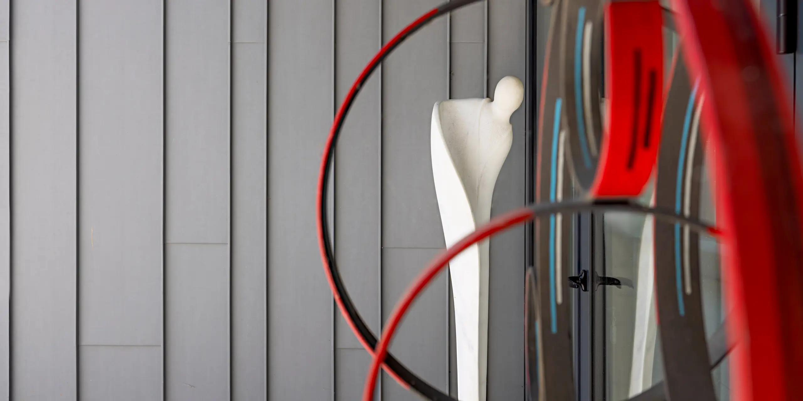 Une sculpture abstraite composée d'anneaux rouges et noirs incurvés encadre une figure humaine blanche, instaurant un dialogue fascinant avec la façade en aluminium PREFA gris quartz de la Scuola Arte e Mestieri F. Bertazzoni de Suzzara. La structure verticale précise des éléments de façade PREFA crée un écrin à la fois serein et élégant, mettant parfaitement en valeur l'installation artistique. La surface mate et veloutée du revêtement en aluminium PREFA souligne la grande qualité et la durabilité exceptionnelle de l'ensemble du bâtiment.